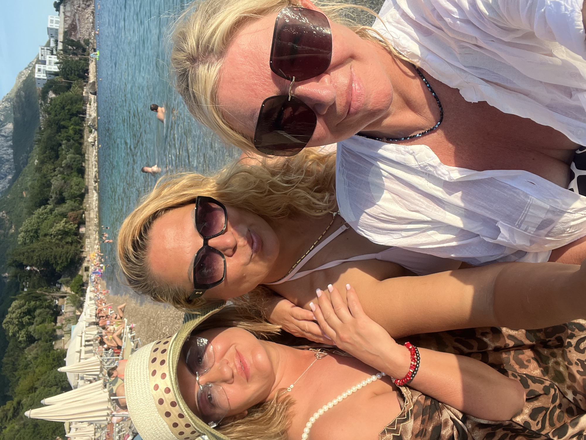 Three women are taking a selfie on a beach in Kamenovo, Montenegro. The woman in the foreground on the right is wearing large, dark sunglasses and a white button-down shirt, unbuttoned to reveal a bikini top. She has blonde hair and is wearing a dark beaded necklace. To her left, another woman with blonde, curly hair is wearing large, dark sunglasses and a bikini top. She is embracing the woman on the right. She is wearing a pearl necklace and a red and black beaded bracelet. The woman on the far left, partially obscured by the other two, is wearing a wide-brimmed straw hat with a polka-dot band and sunglasses. She is wearing a leopard-print dress and a pearl necklace. Behind them, the beach is lined with sun loungers and umbrellas. The sea is calm and blue, with a few people swimming in the distance. Mountains covered in green trees rise in the background, with white buildings nestled on the hillside. The sky is clear and blue. The sun is casting strong shadows, indicating it is either morning or late afternoon.