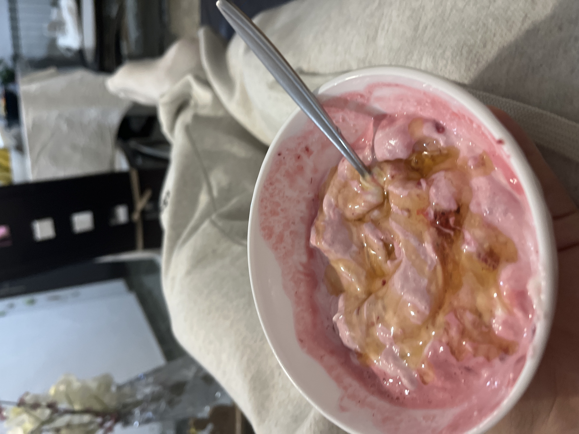 A bowl of pink yogurt with a drizzle of honey is being held, a spoon is inside the bowl. The background is blurred and includes a light-colored garment and a dark-colored object with cutouts. The location is not identifiable as Zlatograd, Bulgaria.