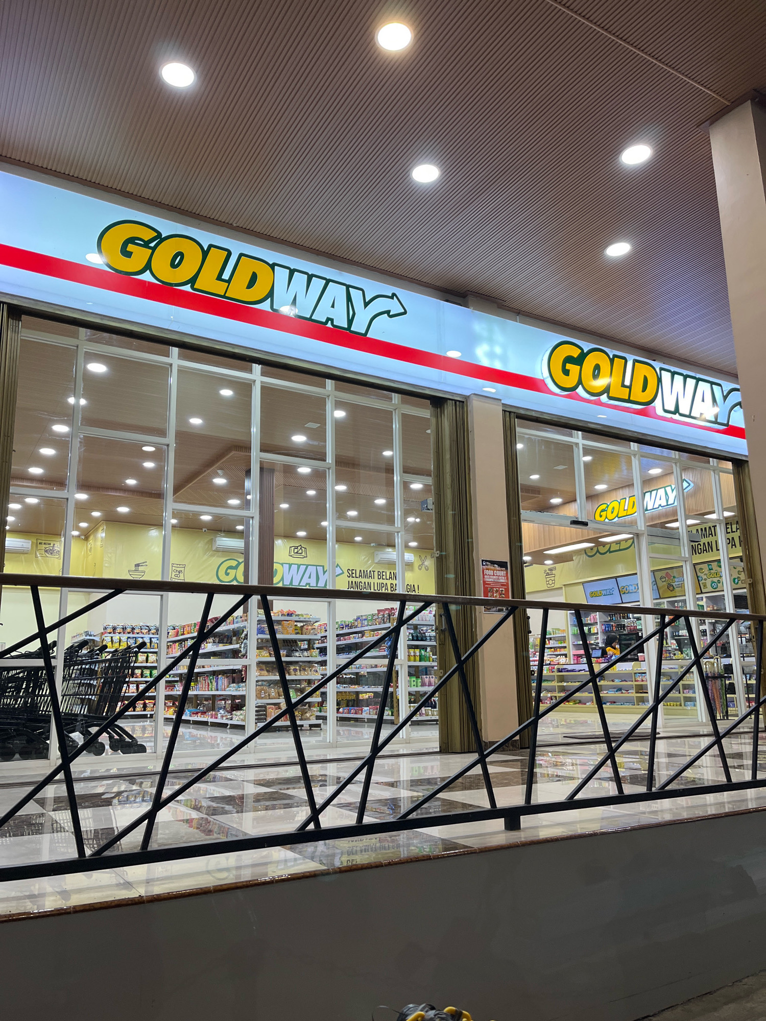 The image shows the exterior of a "GOLDWAY" convenience store in Maja Tengah, Indonesia. The store has a prominent yellow and blue sign with the store name in large, bold letters. The storefront features large glass windows, allowing a view into the brightly lit interior where shelves are stocked with various products. Several shopping carts are visible outside the entrance. The flooring inside appears to be a checkered pattern of light and dark tiles. The scene is illuminated by overhead lights, creating a well-lit ambiance. A black metal railing is in the foreground, partially obscuring the view of the store. The overall impression is that of a modern, well-maintained retail establishment.