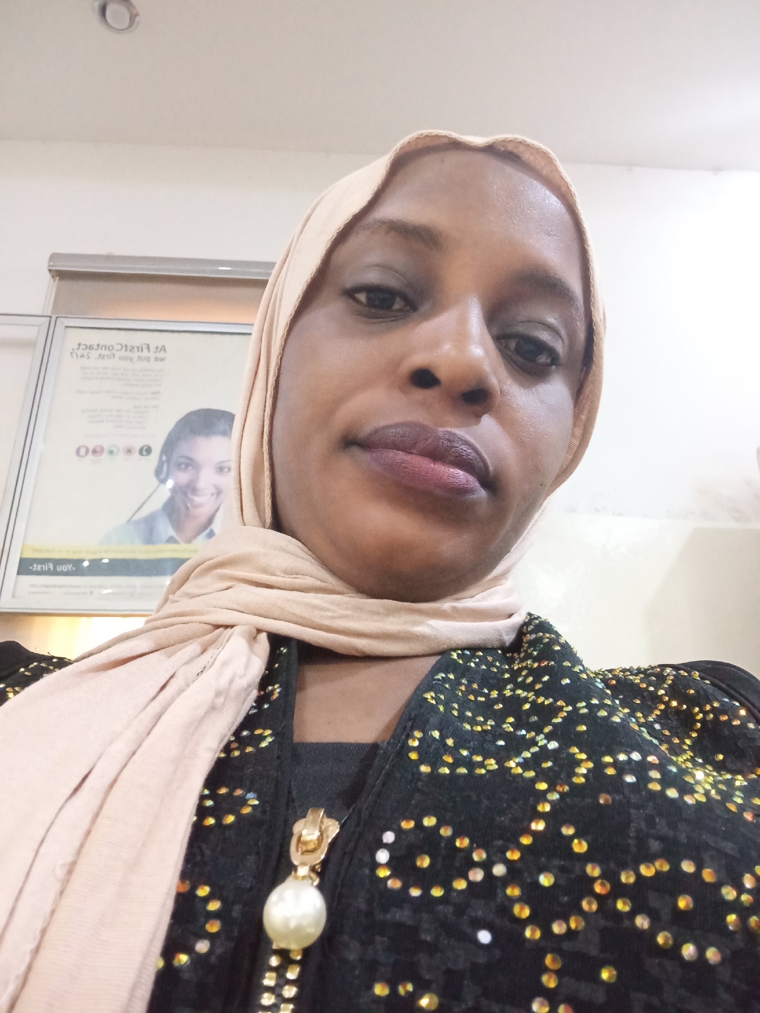 A selfie captures a woman from the chest up, positioned slightly below eye level. She has dark skin and is wearing a light peach or beige hijab that covers her head and neck, draped over her shoulders. Her expression is neutral, and she has dark-toned lipstick on. She is dressed in a black garment heavily embellished with scattered, sparkling yellow and gold sequins or beads that form abstract patterns. A prominent gold zipper pull featuring a large, round, white pearl-like pendant hangs from her clothing.
The background indicates an indoor setting, likely a commercial or public space such as an office or bank. To the left, a framed poster or advertisement is visible, with its text mirrored. Despite the mirroring, key phrases like "A FirsT ConTrAcT" and "me and you put we" can be partially deciphered, suggesting a service-oriented advertisement. The poster prominently features a smiling woman wearing a headset, characteristic of a customer service representative, alongside several circular graphic elements or logos. Above the poster, a portion of a white ceiling and a recessed light fixture are visible. The lighting appears to be artificial indoor illumination. The location is contextualized as Kaduna South, Nigeria.