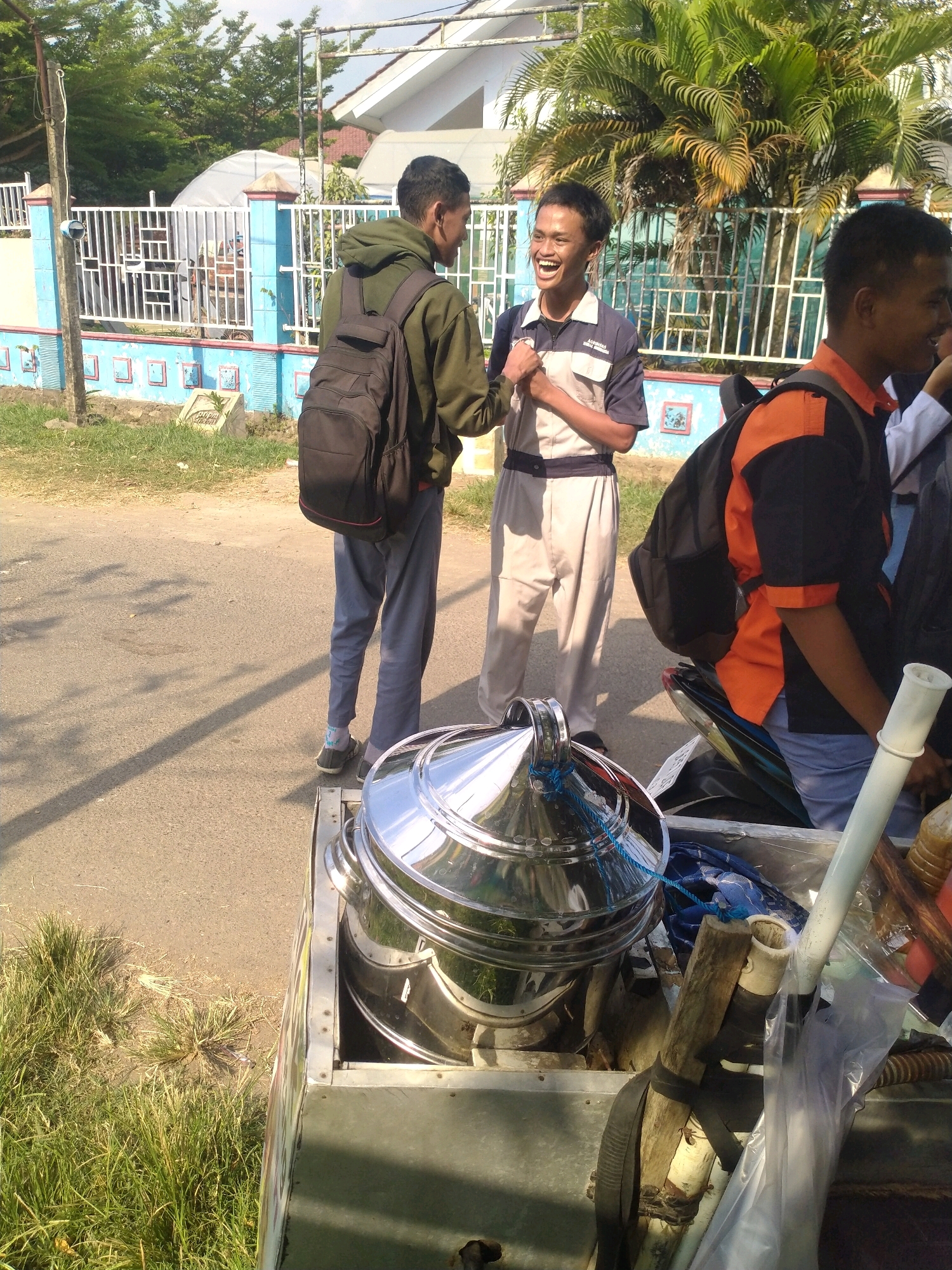 In the city of Begajah, Indonesia, three young men stand on a paved road next to a trailer laden with various items, including a large, stainless steel food steamer and a motor scooter. The man on the left, wearing a green hoodie and black backpack, faces away from the camera, engaged in conversation with the man in the center. The central figure, dressed in a grey and blue collared shirt and grey pants, smiles broadly, his hands clasped together. To the right, another young man in an orange and black shirt, also wearing a backpack, is partially visible. The trailer, positioned in the foreground, contains the shiny steamer, plastic containers, and a roll of blue rope. Behind the men and the trailer, a white metal fence borders a property with lush green trees and a modern building. The scene is illuminated by bright daylight, casting long shadows on the road.