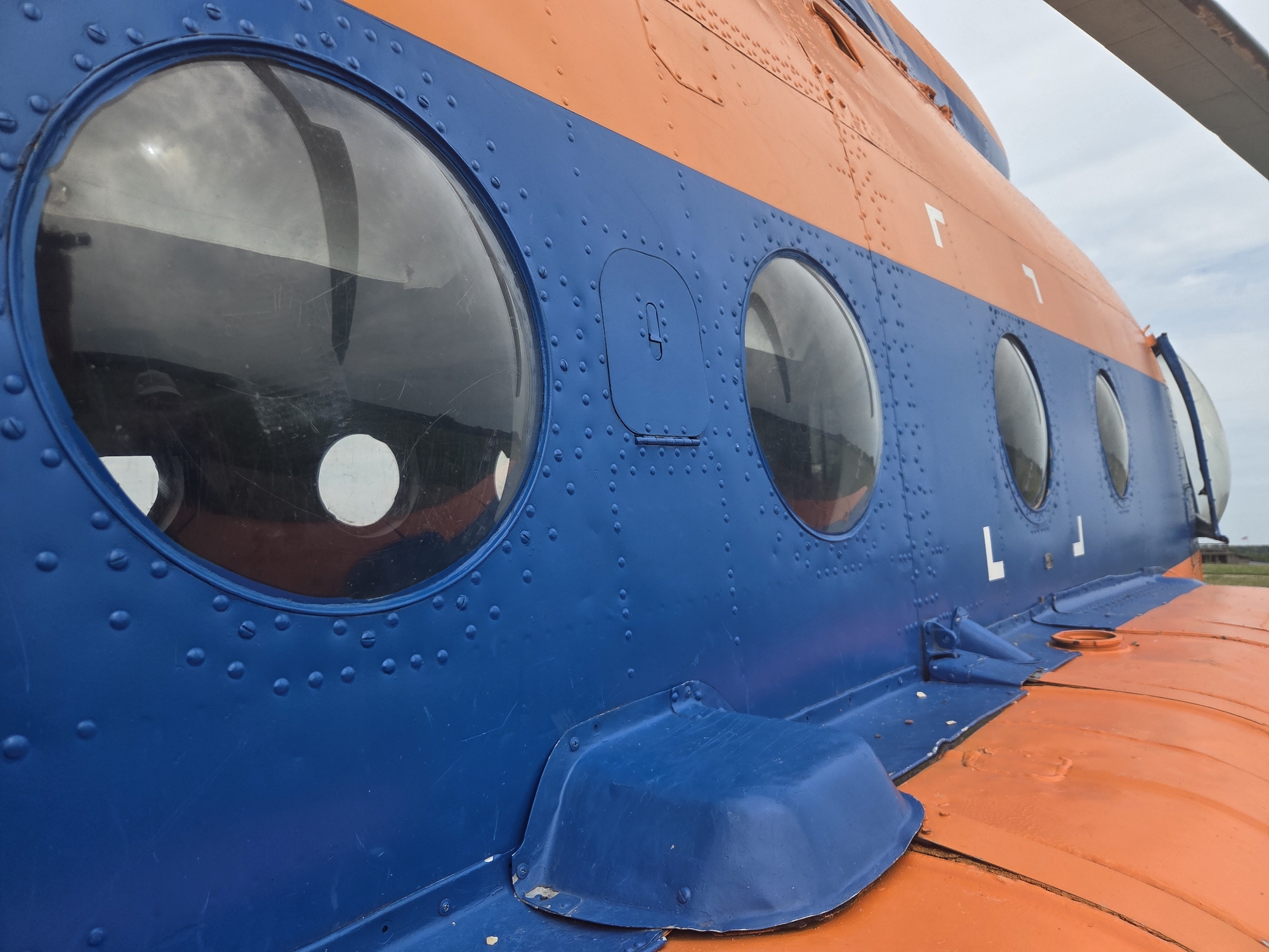 A close-up, low-angle exterior shot shows the blue and orange fuselage of a helicopter parked outdoors. The blue section features several round porthole windows, with the closest one reflecting a cloudy sky and the rotor blades. Rivets are visible along the seam where the blue and orange panels meet, and on the blue panel itself. A small, rectangular access panel with a latch is also present on the blue section. The orange section below is a flat, textured surface. A part of the helicopter's tail boom or a similar appendage extends into the upper right corner of the frame, appearing to be a light gray color. The background is a hazy, light-colored sky with some clouds. The helicopter is situated in an open outdoor area, possibly an airfield or a field, with no visible structures or immediate surrounding cityscape.