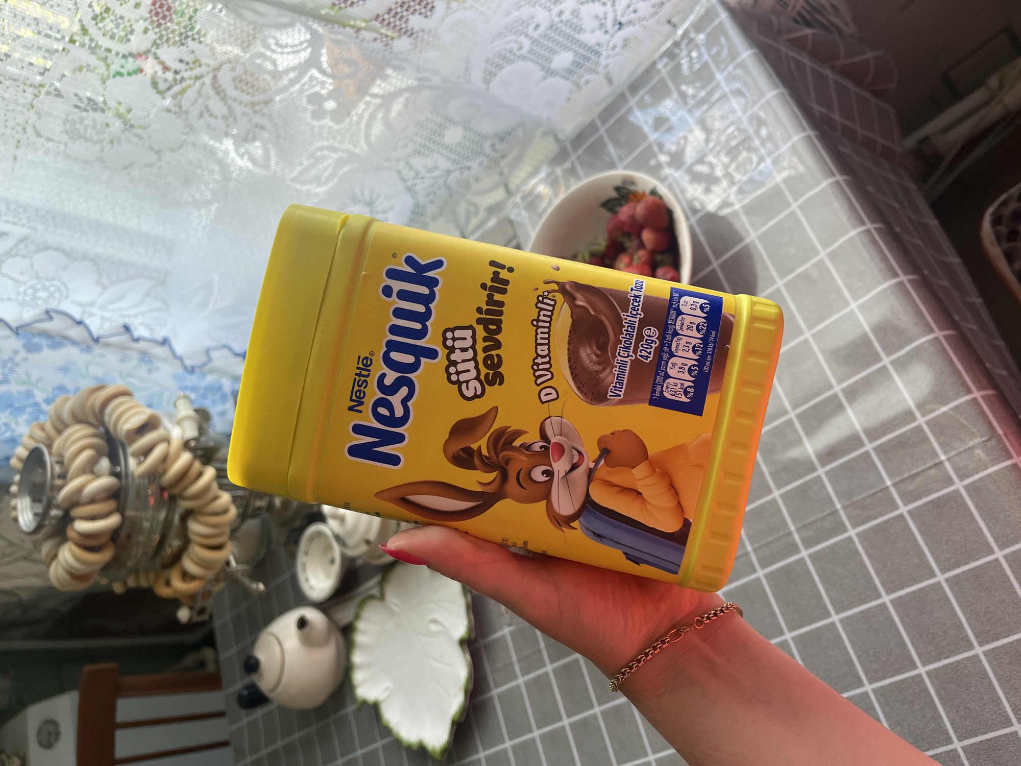 A hand with bright pink nail polish and a delicate gold chain bracelet holds a bright yellow, cylindrical plastic container of Nestlé Nesquik chocolate milk powder. The container is oriented diagonally in the frame, obscuring part of the background. The label on the container is predominantly yellow with blue and brown text and graphics. Prominently displayed is the Nestlé logo and the "Nesquik" brand name. Below this, text in Turkish reads "sütü sevdirir!" which translates to "makes milk lovable!". Further down, there is text mentioning "D Vitaminli" (with Vitamin D) and "Vitaminli Çikolatalı İçecek Tozu" (Vitamin Chocolate Drink Powder), along with nutritional information, including "420ge". A cartoon image of the Nesquik bunny character is featured on the label, appearing to pour chocolate milk from a bag.
The background shows a patterned tablecloth with a grey and white checkered design. To the left of the Nesquik container, a silver decorative object with a coiled, dough-like appearance is visible, possibly a lamp or candle holder. A small white bowl filled with red strawberries is also present in the background. Further to the left and below are other decorative ceramic items, including a white bird-like figure. The lighting suggests it is daytime, with natural light illuminating the scene. The overall impression is a casual, domestic setting, possibly a kitchen or dining area.