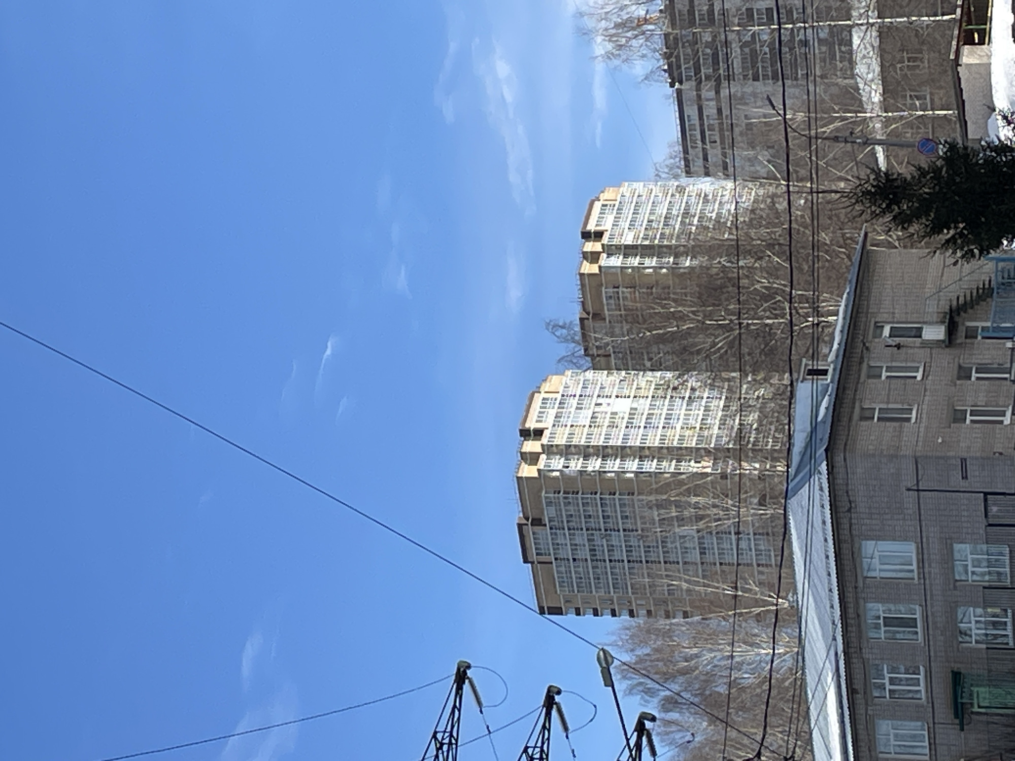 This image depicts an urban landscape in Izhevsk, Russia, under a clear blue sky with minimal, wispy white clouds. The scene is illuminated by daylight, suggesting winter or early spring due to the bare deciduous trees and visible snow.
In the foreground, a two-story grey brick building with a grey corrugated metal roof, partially covered in snow, occupies the lower left. Multiple rectangular windows with white frames are visible on its facade. A small, dark green door or entrance is present on its lower left side. To the right of this building, a dark green coniferous tree stands, its base partially covered in snow. Utility lines and structures crisscross the upper foreground and midground; a large metallic support structure with insulators is visible on the far left, and multiple dark wires stretch diagonally and horizontally across the frame. Snow patches are visible on the ground in the lower right.
The midground features numerous bare deciduous trees with light-colored bark. A lamppost with a light fixture and a blue circular traffic sign is positioned to the right of the coniferous tree. Behind the foreground building, another multi-story building with a grey facade and what appears to be scaffolding or balconies is partially visible on the right.
In the background, two prominent, modern, multi-story residential buildings dominate the skyline. They feature light-colored (beige/tan) facades with numerous windows, some reflecting sunlight, and darker brown upper sections or rooftops. The building on the left is slightly closer to the center. To the far right, another tall, grey multi-story building is partially visible. No people or overt actions are discernible in the scene.