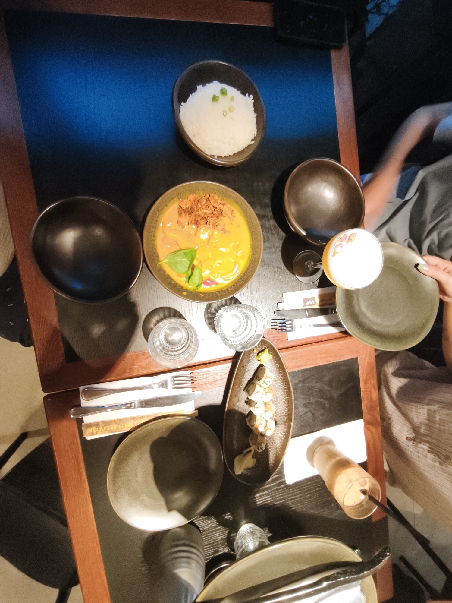 This image captures a table setting at a restaurant in Bnei Dror, Israel, showcasing a meal for at least two people. The table is a dark wood surface with a visible blue tint from lighting, set with various dishes and drinks. In the foreground, a dark, oval-shaped plate is positioned with silverware. To the left, another dark, deep bowl is empty. Above these, a lighter brown, oval dish contains a portion of rice topped with green onions. Adjacent to the rice is another deep bowl holding a vibrant orange curry-like dish garnished with fried shallots and a basil leaf.
To the right of the curry, a tall glass of a light-colored beverage with a frothy top, possibly a cocktail or creamy drink, sits next to a dark, round bowl. Further to the right, a hand with a light-colored sleeve and a green nail is reaching for a flat, greyish-green plate. Below the drinks and curry, a small, elongated dish contains pieces of sushi or similar rolled food items, accompanied by pickled ginger. Two glasses of water are also visible on the table, along with another beverage in a clear glass with an orange liquid. The background is blurred but suggests a dimly lit, indoor environment, possibly a restaurant interior, with hints of people's clothing and arms. The overall impression is of a shared dining experience with a diverse array of food and drinks.