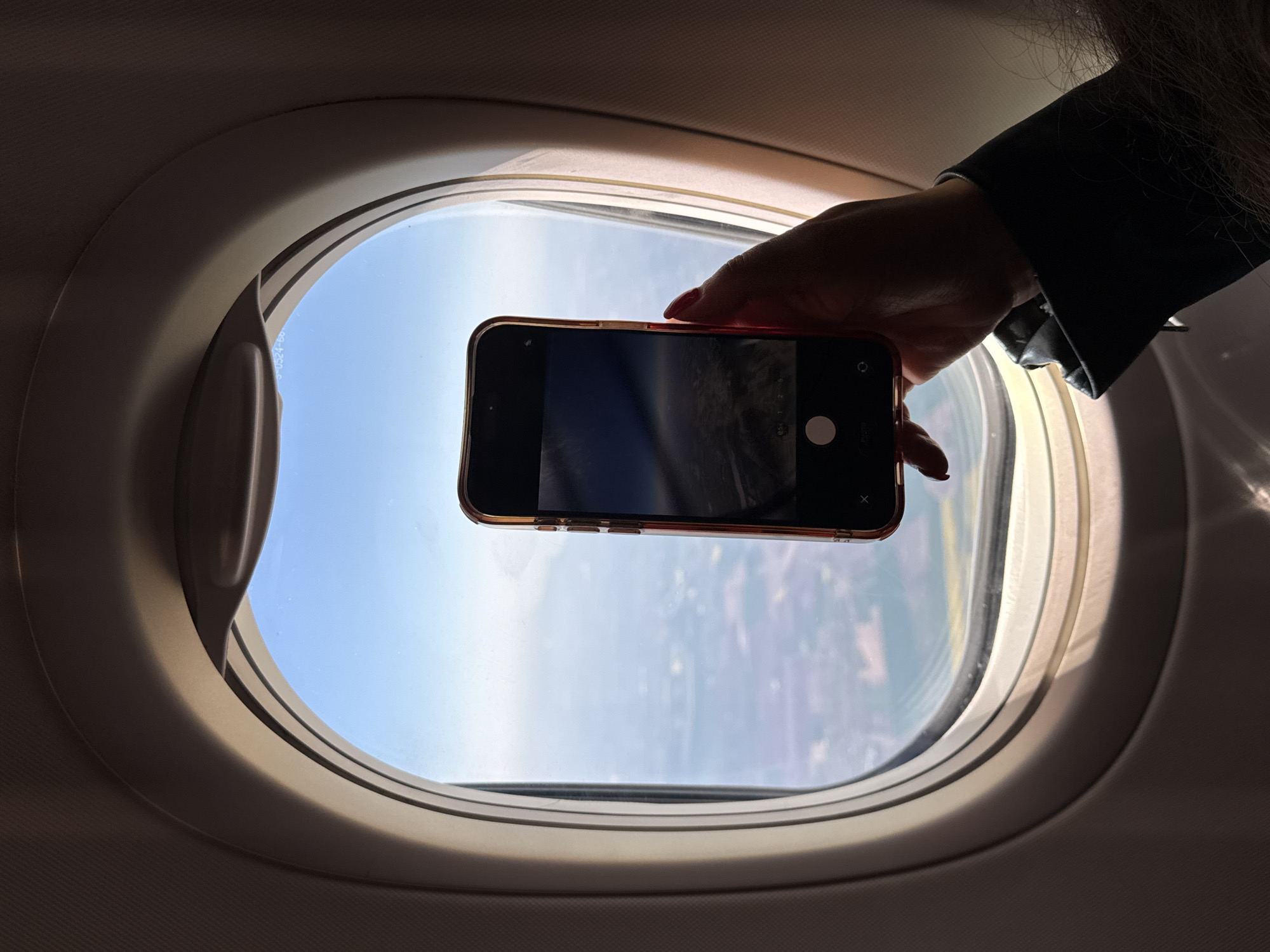 The image presents a close-up, angled view from inside an airplane, centered on an oval-shaped window. A person's hand, with red/pink painted fingernails, is visible on the right, holding a smartphone horizontally up to the window. The smartphone's screen is dark but displays a camera interface, with a white circular shutter button and the word "PHOTO" partially visible on the right side. The phone's casing appears to be a metallic gold or rose gold color.
Outside the airplane window, a bright, clear blue sky is visible, with some distant land or cloud cover creating a softer horizon line below. The internal environment of the airplane, including the window frame and cabin paneling, is significantly darker than the exterior, creating a strong silhouette effect of the hand and phone. This contrast emphasizes the act of capturing the illuminated view from the shadowed cabin. The time of day is clearly bright daylight. Faint text, "S-0524-60", is partially visible on the bottom edge of the window frame. The scene captures the common activity of passengers documenting their flight experience. No specific landmarks are visible to indicate the given location context of Sofia, Bulgaria.