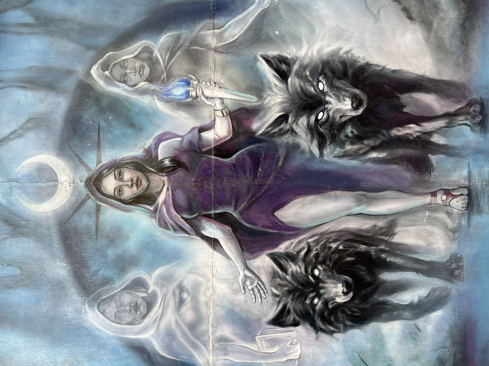 The image shows a mural with an allegorical scene. A woman wearing a purple dress is centrally positioned, holding a torch with a blue flame. A crescent moon and star-like spikes frame her head. Above and below her are ethereal figures, appearing as ghost-like women in hooded cloaks. Two wolves flank the central woman, positioned to her right. The background is a blue and white blend, suggestive of a celestial or otherworldly setting. The artwork appears to be weathered. The mural is located in the city of Ufa, Russia.