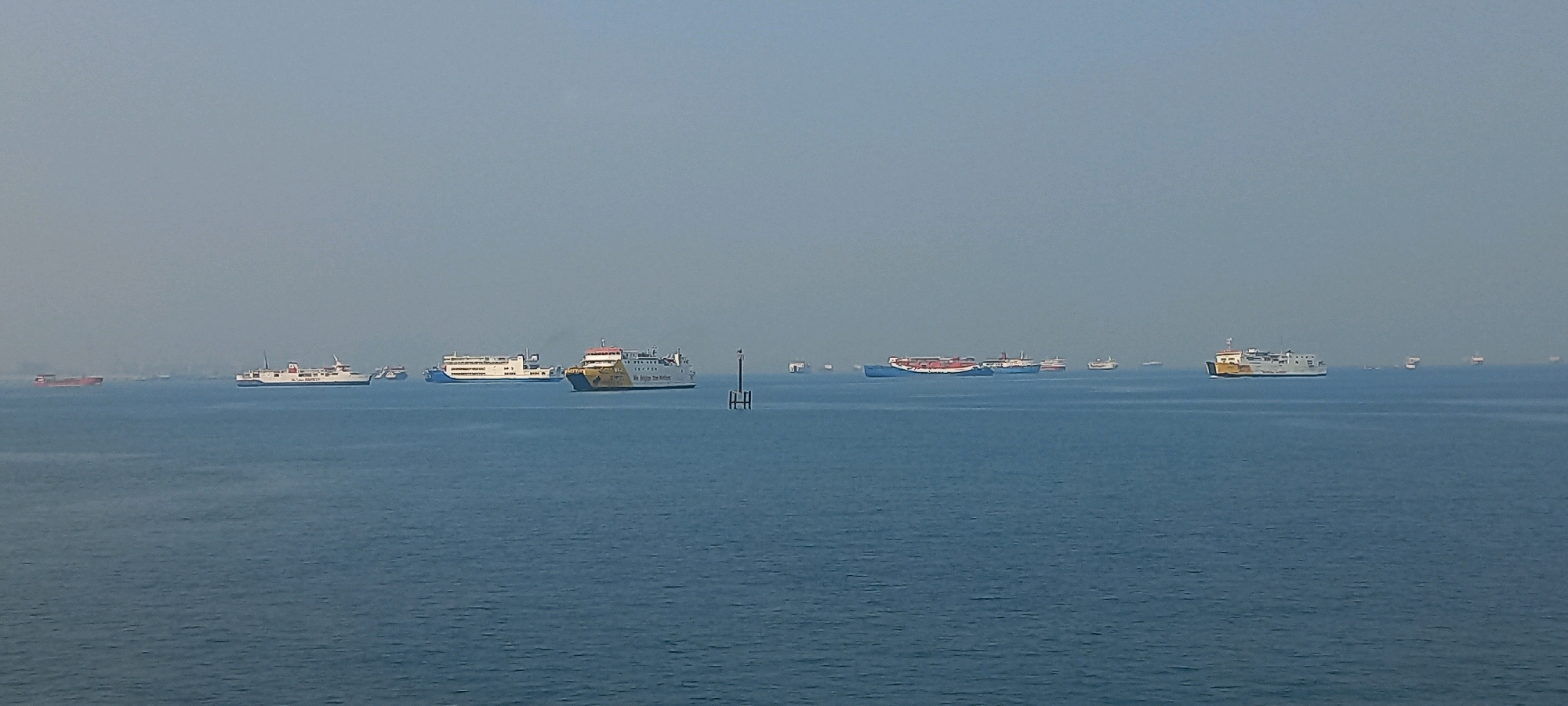 The image depicts a wide expanse of calm, blue sea under a hazy, pale blue sky, likely taken from a high vantage point in or near the city of Cilegon, Indonesia, given the presence of numerous vessels. A variety of cargo ships and ferries are visible in the distance, some appearing as specks on the horizon, while others are more clearly defined.
In the mid-ground, several larger vessels are more prominent. To the left, a white ferry with a blue hull is moving away from the viewer. Slightly to its right and closer to the center, a white ferry with a distinctive yellow and black hull, possibly a passenger or car ferry, is also visible. Further to the right, a long, blue cargo ship with red containers stacked on its deck is positioned horizontally. Behind it, other vessels of varying sizes can be seen. On the far right, a smaller, white vessel appears to be a ferry or a similar type of ship.
A solitary, dark gray navigational buoy with a visible structure stands in the middle right of the frame, marking the water. The overall scene suggests a busy maritime area, common in Cilegon, known for its port and industrial activity. There are no people visible in the image.