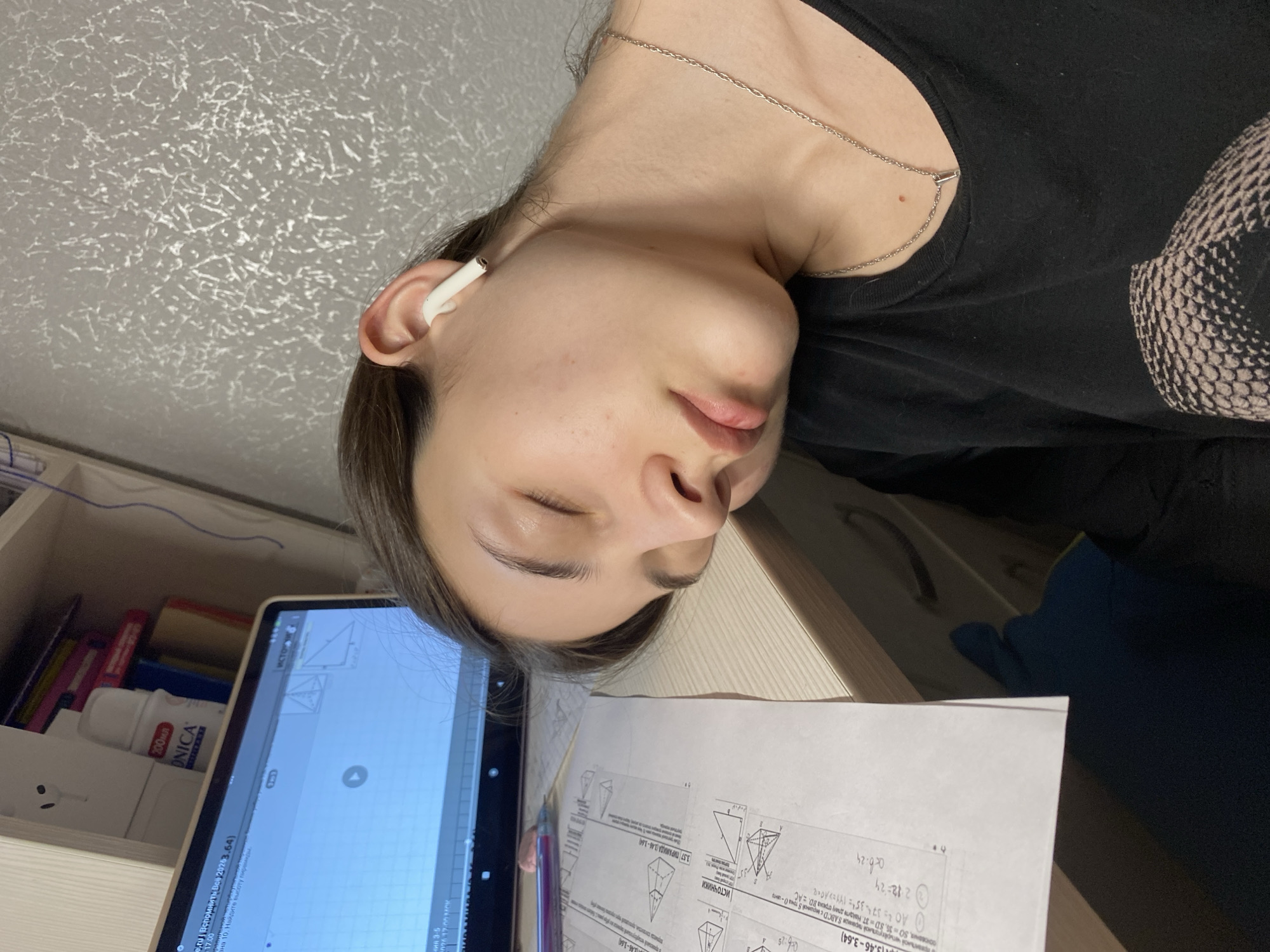 A young woman is dozing at a desk. She is wearing a black top, a silver necklace, and a white wireless earbud. Her eyes are closed. Papers with mathematical diagrams and equations are on the desk, next to a laptop displaying a graphic of geometric shapes. Books and a white container with the word "ONICA" are visible in the background, suggesting an indoor setting. The location is not possible to determine from the image.