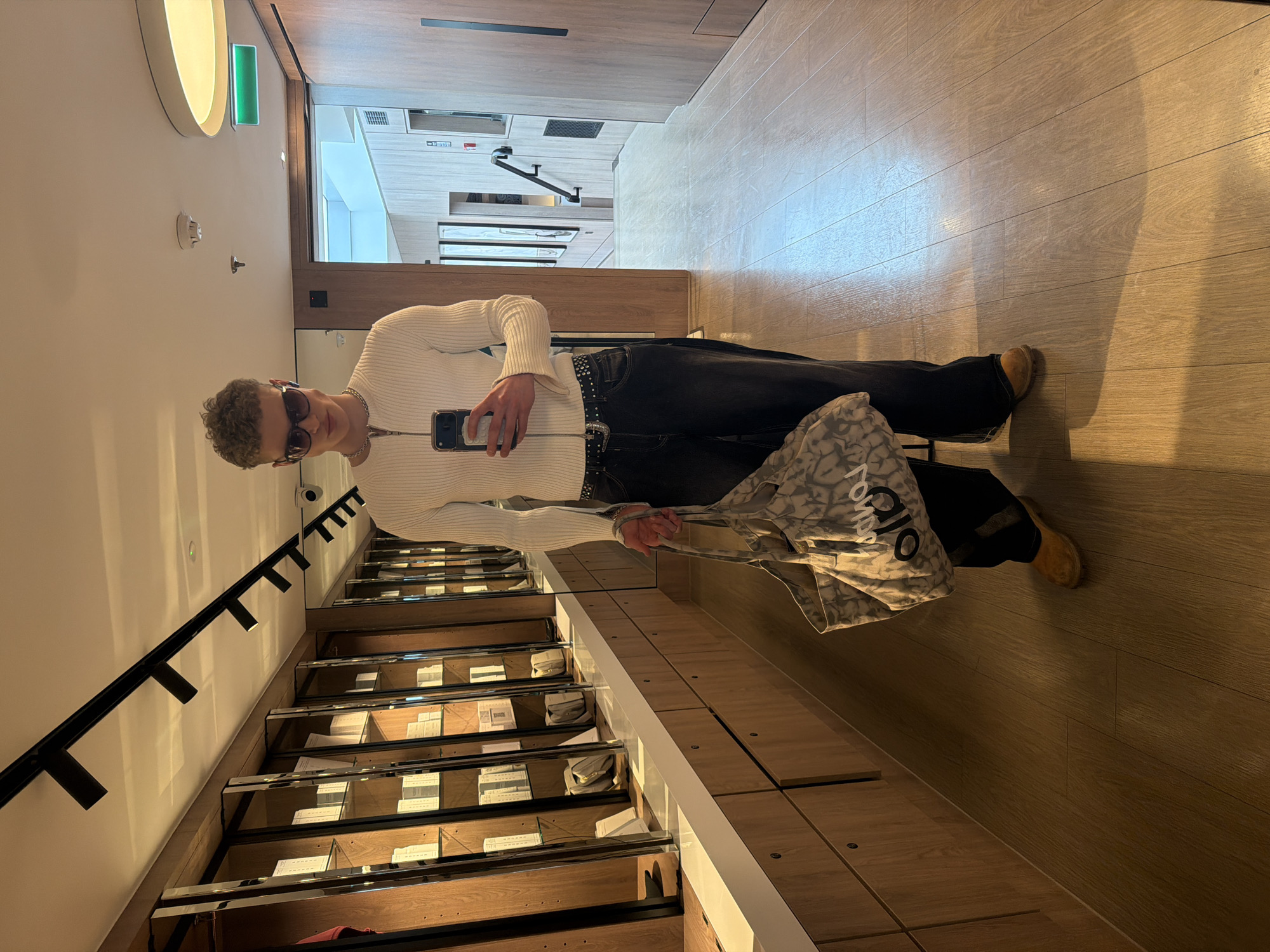 A man is posing in what appears to be a clothing store in London, United Kingdom, as evidenced by the "ALO" tote bag he is holding. The man is wearing sunglasses, a white, ribbed long-sleeved shirt, black jeans with a studded belt, and tan shoes. He is holding a cell phone. He is standing next to a retail display rack filled with folded items. The store's interior includes wood flooring and recessed lighting.