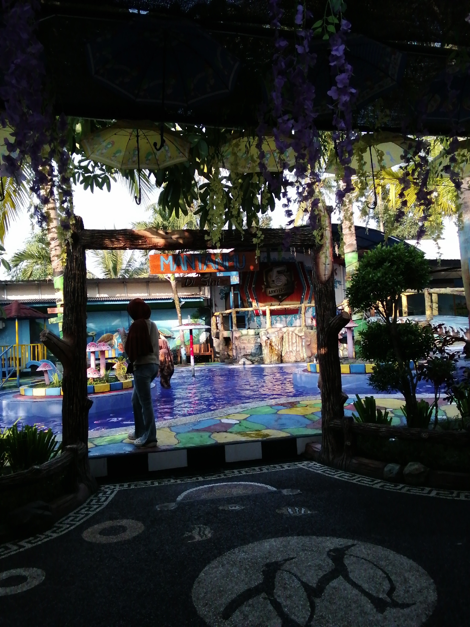 This is a full-shot, outdoor photograph taken during the daytime, likely in Kebonsari, Indonesia, depicting an entrance to a water park or recreational area. The foreground shows a dark, pebbled walkway with decorative circular patterns. Framing the view is a wooden archway adorned with artificial purple and yellow hanging flowers.
Through the archway, a person wearing jeans, a brown headscarf, and a light-colored shirt is standing, facing away from the camera. They are holding a brown backpack. To the right of the pool area, a small, green bush and a larger, leafy tree are visible.
The background reveals a colorful swimming pool with blue water and a mosaic-tiled bottom featuring various shapes and colors. Surrounding the pool are structures with colorful walls and roofs, including what appears to be a stage or activity area with a large sign that reads "MANTA..." and "5 ANNIVERSARY." There are also playground-like elements with mushroom-shaped decorations and a turtle statue. Palm trees and other greenery are visible in the distance, suggesting a tropical or subtropical climate. Umbrellas are suspended from the ceiling structure in the foreground.