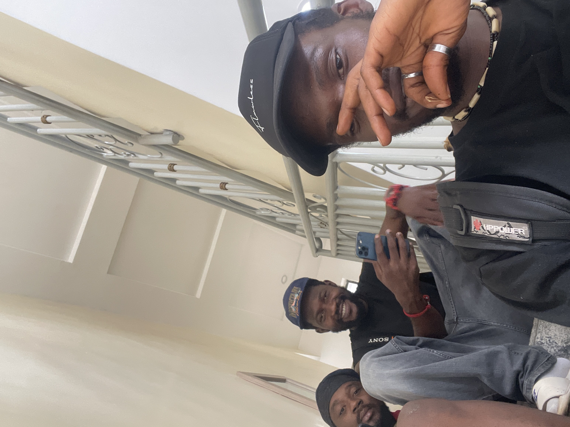A low-angle, tilted photograph captures three dark-skinned men in what appears to be an indoor or semi-enclosed setting, likely in Gwarinpa, Nigeria, indicated by the architecture and casual attire. The area is brightly lit, with light beige walls, a white ceiling, and a white metal railing suggesting a stairwell or corridor.
In the foreground, a man wears a black baseball cap with "Flawless" written in white script, a black t-shirt, and a necklace of black and light-colored beads. He has silver rings on his index and ring fingers, and his hand partially obscures his face as he looks towards the camera.
Behind him, a second man smiles at the camera, holding a blue smartphone. He wears a blue baseball cap, a black t-shirt with "SONY" in white lettering, grey jeans, and a red bracelet. A black shoulder bag featuring a white and red "HPPOWER" logo is visible.
Further back, a third man, wearing a black beanie or cap and a dark t-shirt, also looks towards the camera. He wears light-colored sneakers. The men appear relaxed and are posing casually, possibly for a selfie.