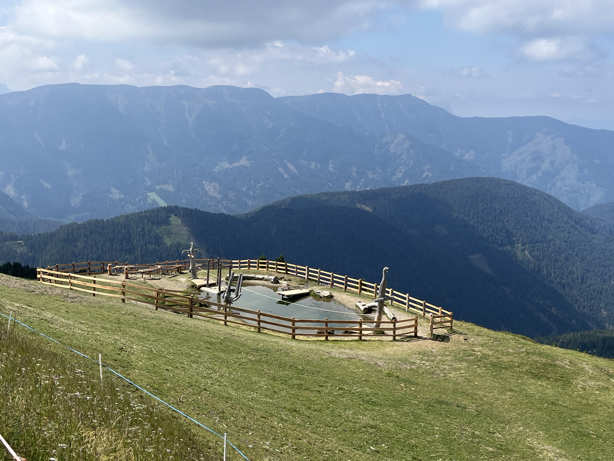 This image depicts a scenic mountain landscape with a man-made water feature and seating area enclosed by a wooden fence. The area is situated on a grassy slope with wildflowers in the foreground. In the background, layers of blue-toned mountains recede into the distance under a cloudy sky. There are no people or animals visible in the frame. The water feature appears to be a pond with some wooden elements, possibly for play or decoration. The overall impression is one of a peaceful, natural environment with a touch of human intervention for recreational purposes. The location is not specified as Brixen - Bressanone, Italy, but rather a mountainous region.