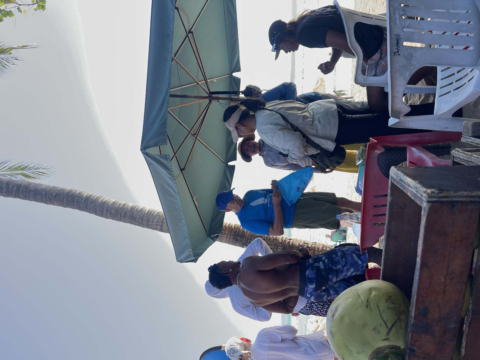 A group of people are gathered on a beach in Kuta, Indonesia, under a large green umbrella. The scene takes place outdoors on a sunny day, with a palm tree trunk visible behind the umbrella. A man in a blue t-shirt and hat is holding a blue plastic bag. Several other individuals are standing and talking. In the foreground, a wooden crate holds coconuts, and plastic chairs are scattered on the sand. The ocean is visible in the background.