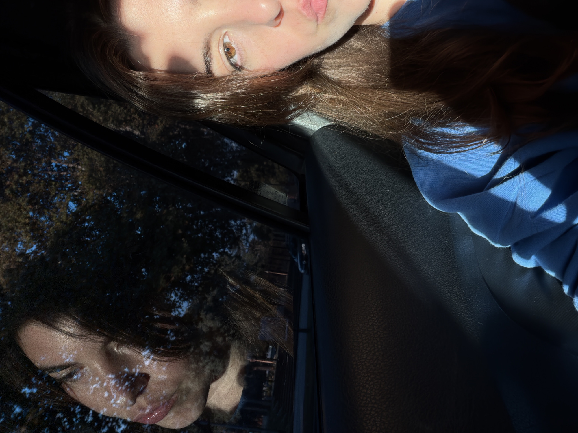 This media file captures a close-up portrait of a young person inside what appears to be a car, notable for its strong natural lighting and a prominent reflection. The image is rotated 90 degrees clockwise from an upright perspective.
The primary subject is a young individual with fair skin, expressive brown eyes, and long, straight, rich brown hair. They are wearing a light blue long-sleeved garment, possibly a sweatshirt. Direct sunlight strongly illuminates parts of their face, including the forehead, bridge of the nose, cheek, and lips, as well as sections of their hair, creating distinct highlights and shadows. Their expression is serene and contemplative.
A clear reflection of the person's face and upper body is visible in the car window on the left side of the frame (when mentally rotated upright). This reflection shows intricate patterns of light and shadow, likely caused by sunlight filtering through foliage outside, cast across their face.
The setting appears to be inside a vehicle. A dark, textured surface, possibly a car seat or interior panel, is visible on the right side of the image. Through the car window and within the reflection, dark silhouettes of trees or dense vegetation and patches of blue sky are discernible, indicating an outdoor environment.
The strong, directional sunlight suggests the photo was taken during either the morning or late afternoon. The lighting creates a high-contrast scene with sharp shadows. No legible text is visible within the image; although some indistinct, pixelated shapes are present in the distant background of the reflection, they are unreadable. The location context of Buenos Aires, Argentina, is provided, though no specific visual cues within the image confirm this location.