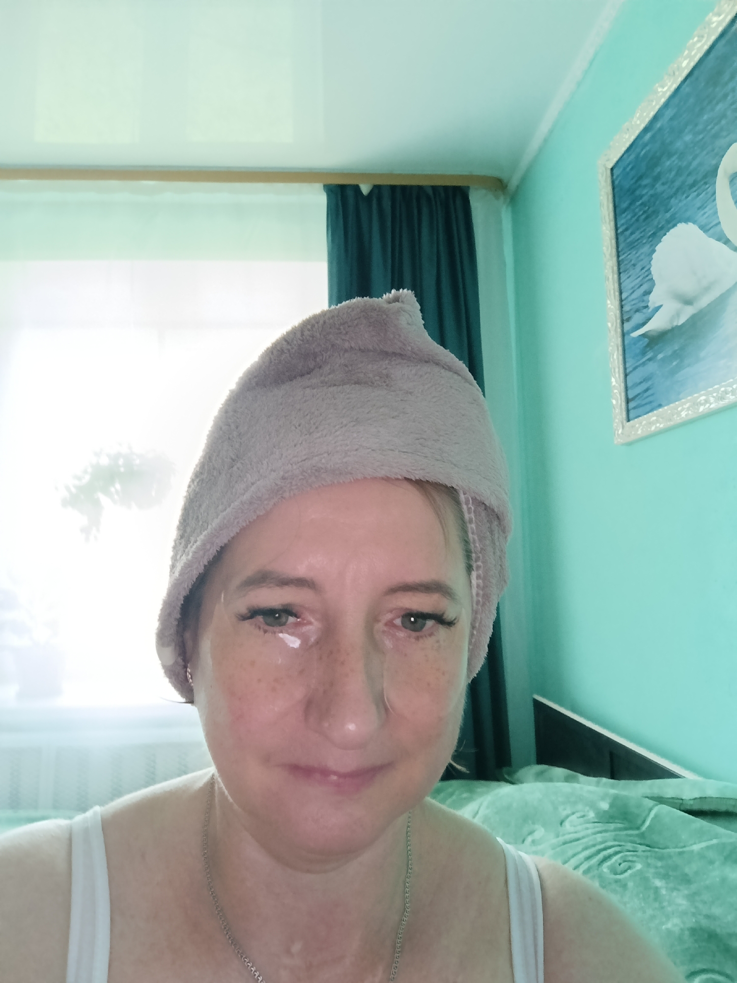 A woman with freckles and a gray towel on her head is looking at the camera. She is wearing a white tank top and a silver necklace. There are eye patches under her eyes. The background shows a window with sheer curtains, a teal wall, and a framed picture of a swan. The room appears to be a bedroom, possibly in Tugulym, Russia.