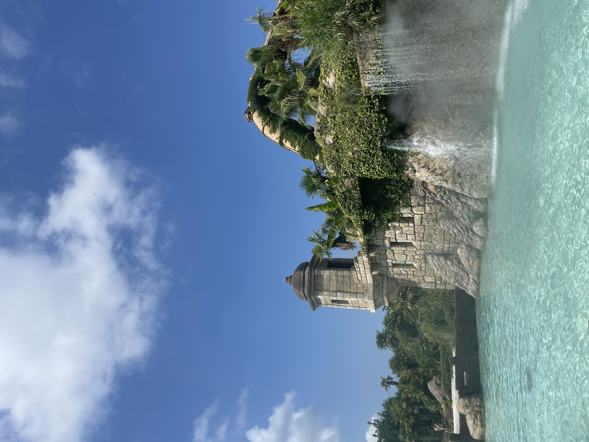 The image depicts an outdoor water park scene in Jesolo, Italy, on a sunny day with a clear blue sky and scattered white clouds. The foreground features a crystal-clear turquoise pool of water, with sunlight shimmering on its surface. To the right, a waterfall cascades down a rocky, vine-covered cliff face, creating a misty spray. In the mid-ground, a stone tower with a domed roof and arched windows stands on a rocky outcrop, reminiscent of a medieval fort. Lush green vegetation, including palm trees and ivy, covers the structures and surrounding cliffs, adding to a tropical ambiance. In the background, more palm trees and dense foliage are visible. A person is partially visible standing on a sandy area to the far left. The overall impression is one of a resort destination designed to evoke a sense of adventure and relaxation.