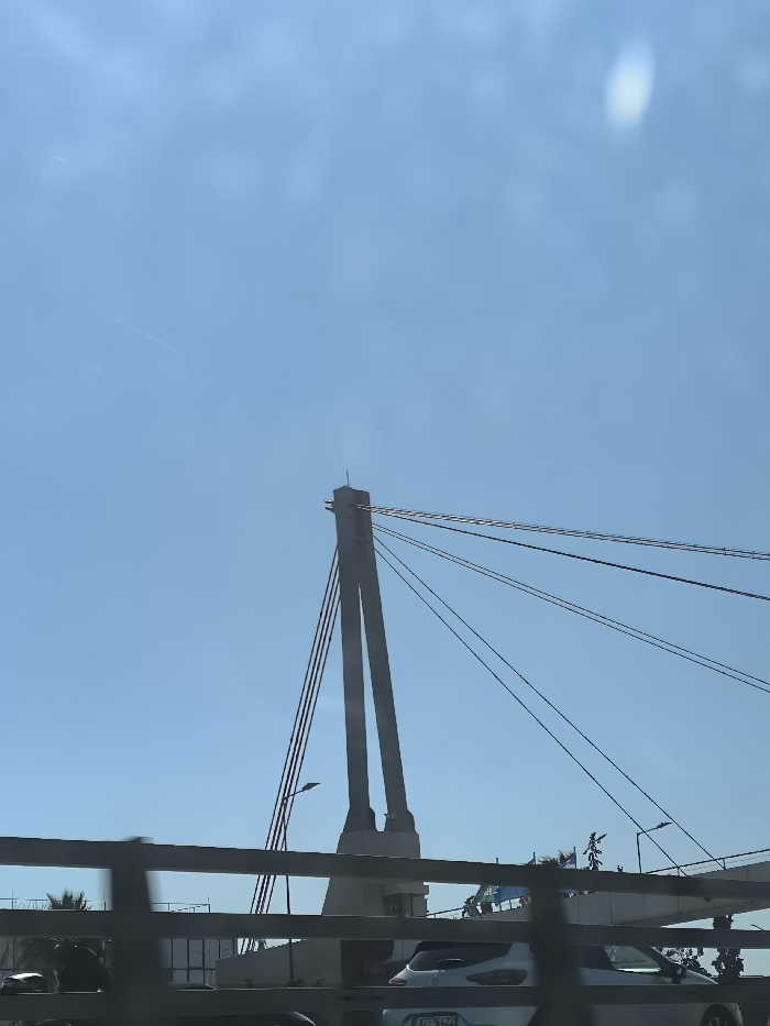 The image shows a portion of a cable-stayed bridge against a clear blue sky with a few white clouds. The image is taken from the perspective of a vehicle driving on the bridge, looking towards the front of the vehicle. The bridge is made of concrete and steel, with a series of cables connecting the bridge deck to the towers. The image is focused on the tower and cables to the right side of the bridge. There are other vehicles on the bridge, and trees and buildings can be seen in the distance. The image is captured under a bright sun. The location is the *Charilaou Trikoupis* Bridge in Egaleo, Greece (coordinates: 37.979, 23.679).