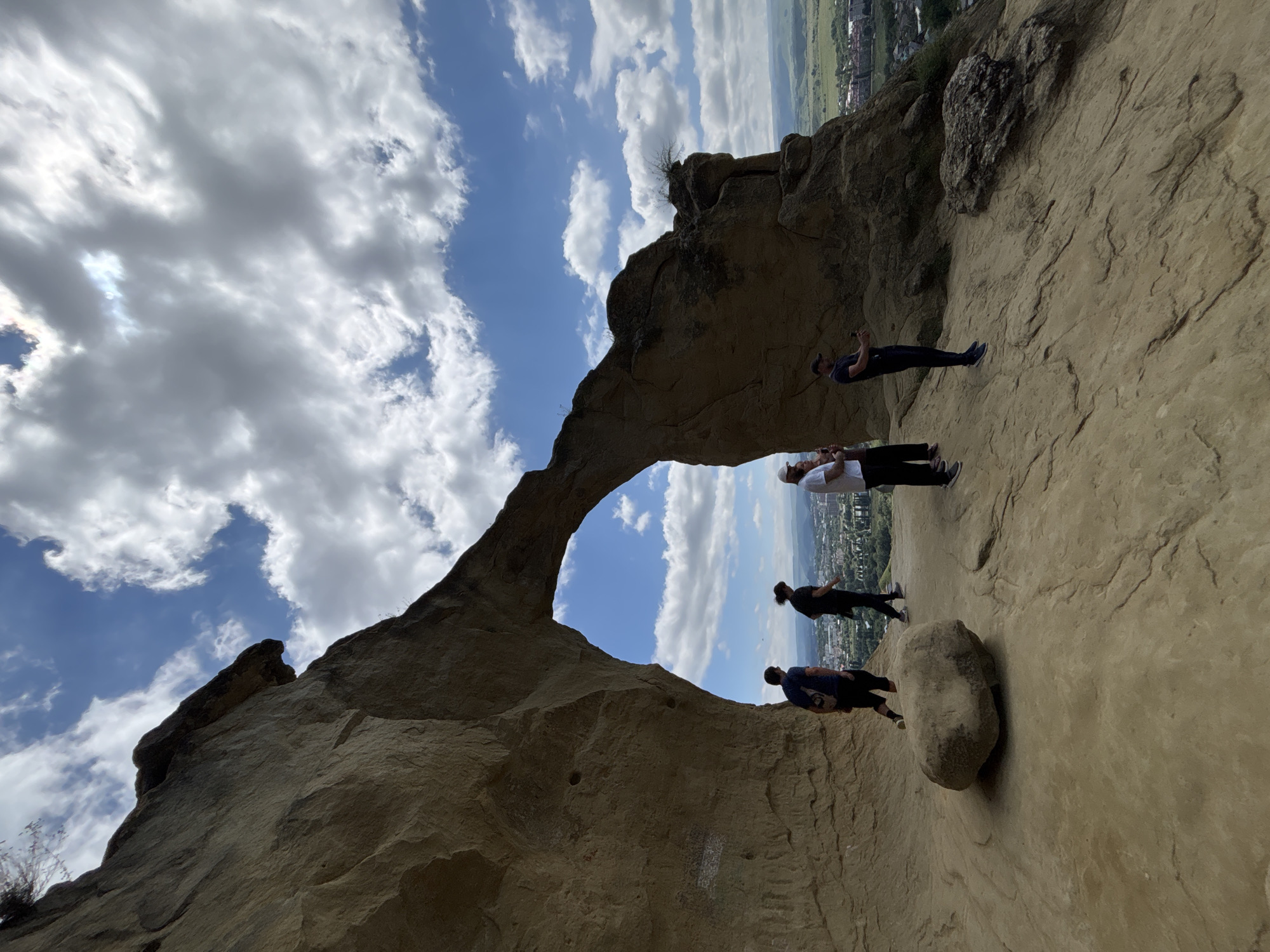 An outdoor, eye-level, wide-angle shot captures a natural rock arch framing a distant view of a city nestled within rolling hills under a partly cloudy sky. The rock formation is a prominent, weathered tan-colored structure with a large opening resembling an arch. Within this archway, a group of four individuals are visible.
On the left side of the arch, standing on the rocky ground, is a person wearing a dark blue t-shirt and dark pants, with a backpack. To their right, a person in a black t-shirt and black pants stands looking down. Further to the right, inside the arch, a person in a white t-shirt and dark pants is holding a phone, possibly taking a photo. Next to them, a person in a dark blue t-shirt and dark pants is also holding a phone and appears to be taking a picture.
Outside the arch, to the right, the ground slopes down, revealing a city with buildings and greenery. The sky is a vibrant blue with scattered white and grey clouds. A large, rounded rock sits prominently on the ground to the left of the arch. The overall scene suggests a tourist destination or a scenic overlook. The context provided indicates the location is in the city of Mirny, Russia.