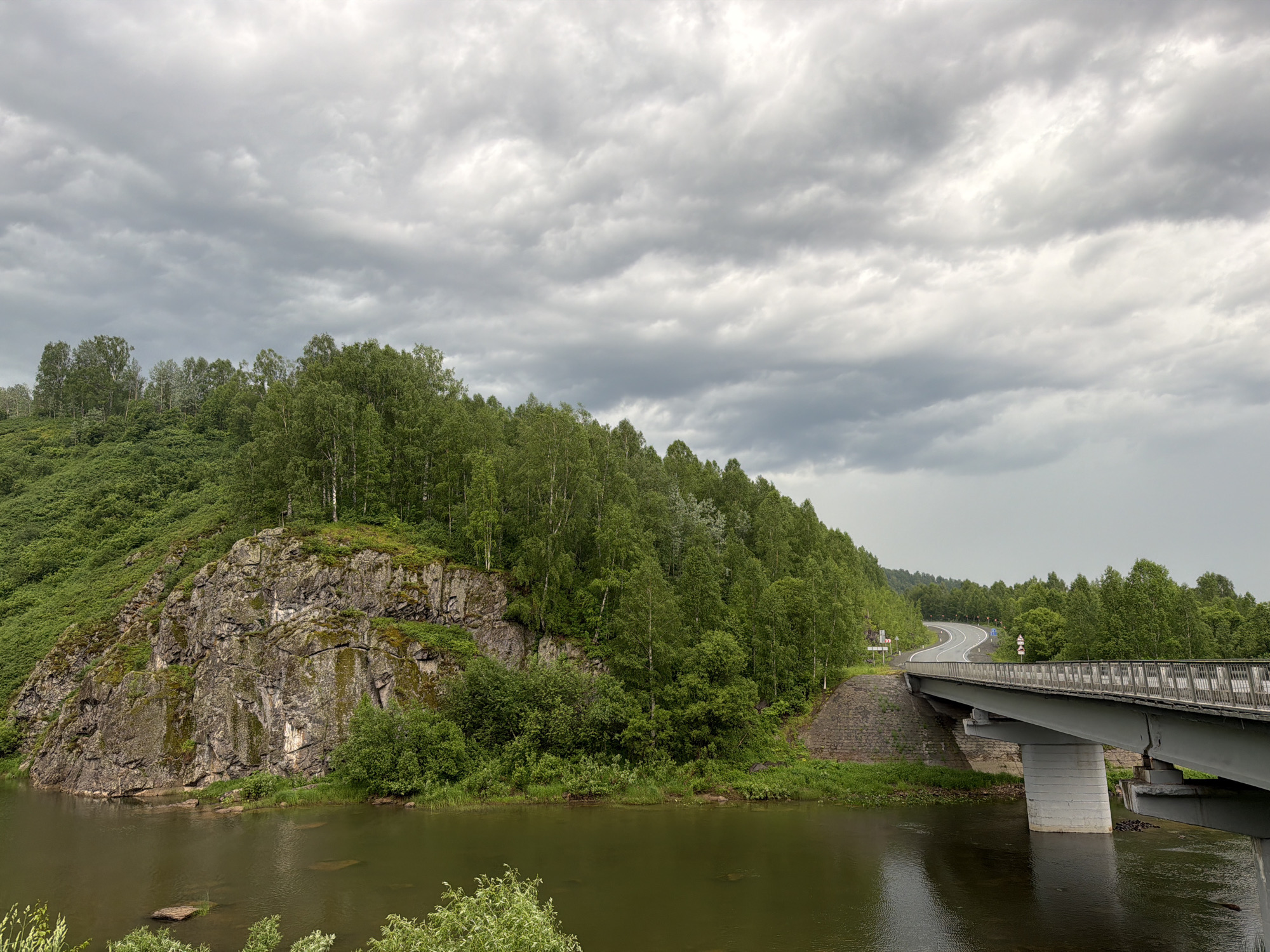 A wide-angle, eye-level shot captures a scenic view of a river flowing through a lush, green landscape under a cloudy sky in Mundybash, Russia. On the left, a steep, rocky cliff face covered in moss and vegetation rises from the riverbank, leading to a dense forest of deciduous trees that stretches across the hillside. A modern concrete bridge with metal railings spans the river on the right, carrying a winding asphalt road that disappears into the distant treeline. The water in the river is dark and reflects the overcast sky. The overall atmosphere is serene and natural, suggesting a quiet, rural setting.