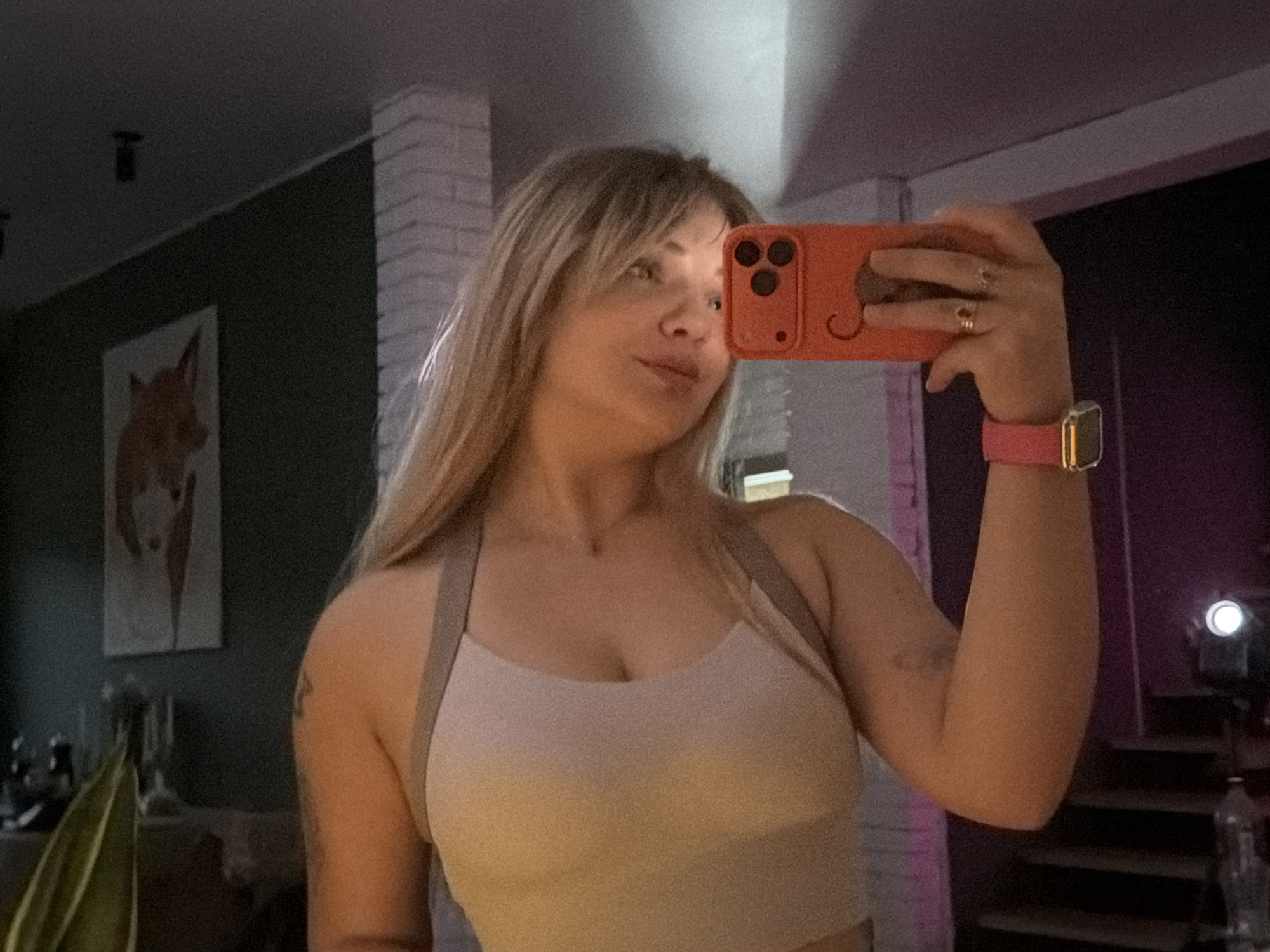 A young woman takes a selfie using an orange-cased iPhone in what appears to be her home. She has long blonde hair with bangs and is wearing a light-colored athletic top with straps. A pink Apple Watch is visible on her left wrist. The background includes a dark green wall with a framed painting of a fox and a white brick pillar. A staircase and a light source are visible in the dimly lit area behind her. The location is Большая Шильна, Russia, based on metadata.