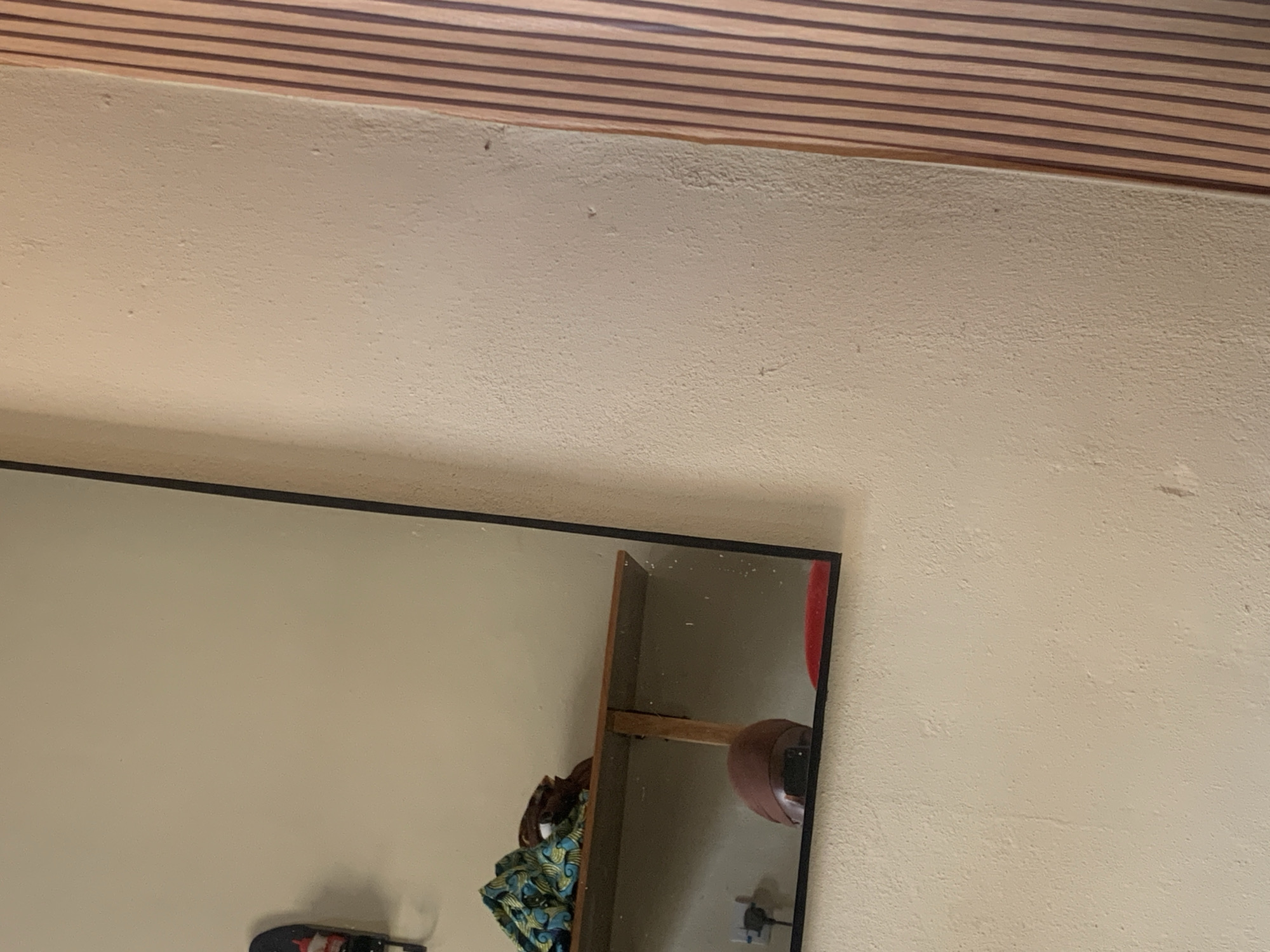 The image shows a close-up view of a wall with a mirror reflecting a shelf with some items. The wall is painted a light, textured color. To the right of the wall, there's a section of vertical wooden slats, creating a striped pattern.
In the reflection of the mirror, a wooden shelf is visible, supported by a wooden bracket. On the shelf, there are two items: a bundle of fabric with a blue, green, and yellow pattern, and a brown object that appears to be part of some clothing or material. Below the shelf, in the reflection, a brown, dome-shaped object is visible, possibly a hat or a similar accessory. A red object is also partially visible in the bottom of the mirror's reflection. In the background, a black electrical outlet is seen on the wall.
The scene is indoors, likely in a residential or storage space. The lighting is ambient, creating soft shadows. The image is taken from a medium distance, focusing on the wall and the reflection in the mirror. There are no people visible in the image. The overall impression is one of a simple, uncluttered space.