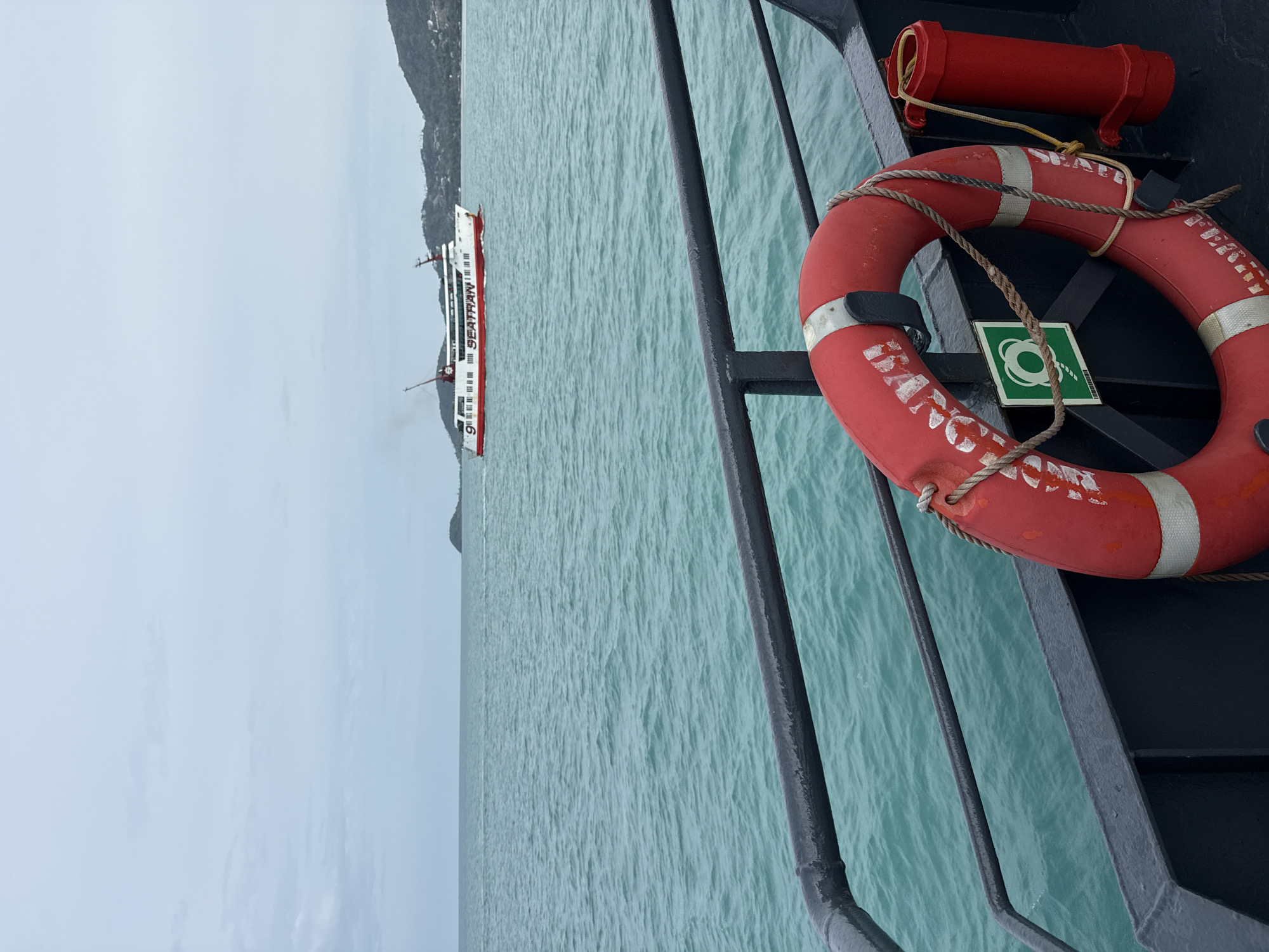 The image shows a view from the deck of a ferry in the Gulf of Thailand, likely near Ko Samui District, Thailand. In the foreground, a red life preserver is attached to a railing. The sea is a vibrant turquoise and appears slightly choppy. In the middle ground, a white and red passenger ferry with the name "SEATRAN" emblazoned on its side is sailing on the water. In the background, a lush, green island, characteristic of Ko Samui's landscape, is visible. The sky is overcast with a soft, diffused light. The overall scene depicts a typical ferry journey in the waters surrounding the popular tourist island.