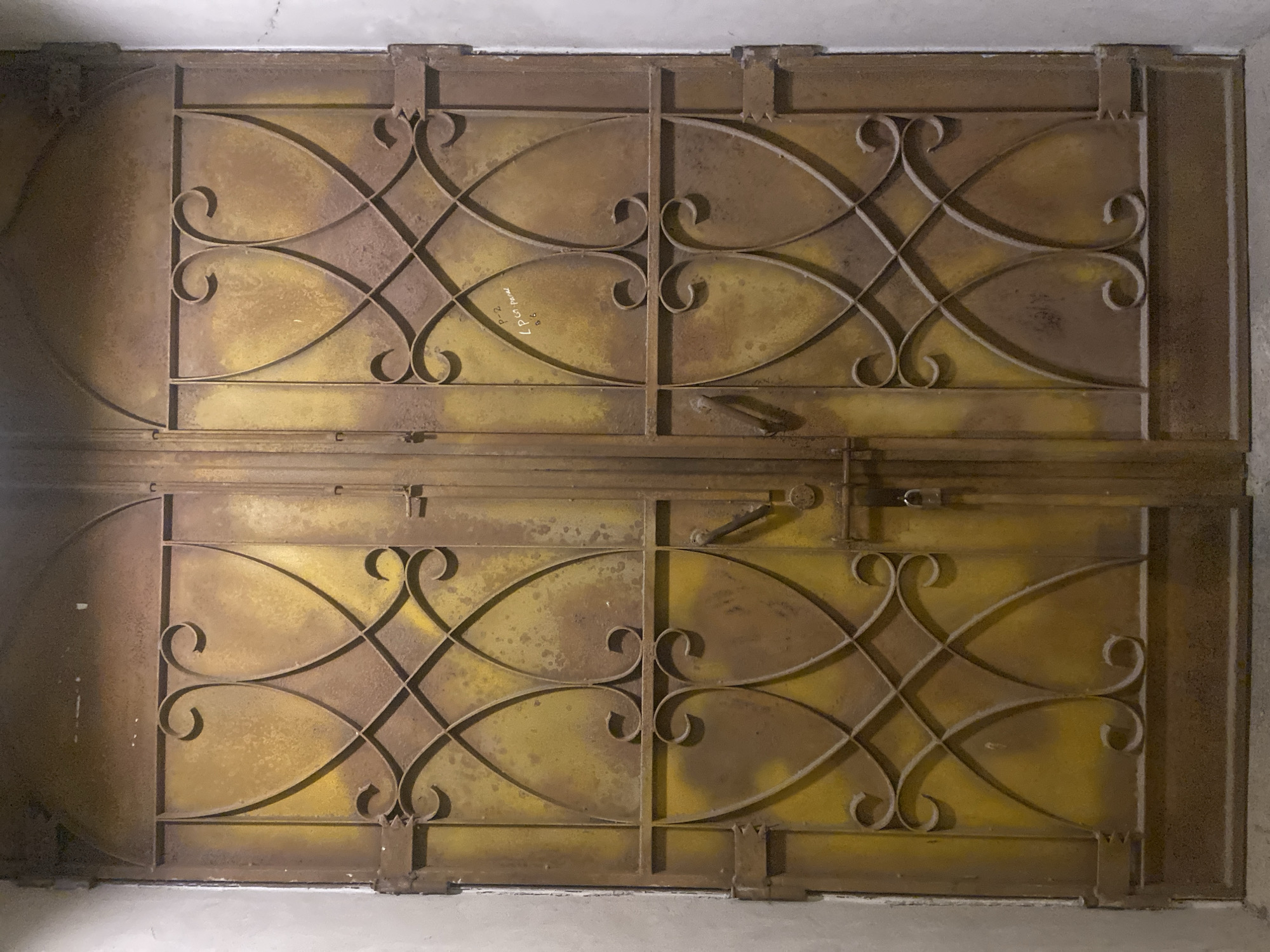 Two weathered, yellow metal double doors with ornate wrought iron detailing are situated within a recessed entryway. The doors are marked with rust and wear, indicating age and exposure. The left door has two visible handles and a lock. A padlock is attached to the left door. The right door also has one visible handle. The doors are framed by a plain, light-colored wall on either side. A section of the right door has handwritten text in black marker, reading "P-2 Lpgn. Parall". The floor in front of the doors appears to be concrete. The scene is lit with artificial light, creating some glare and shadows on the textured surfaces of the doors and walls.