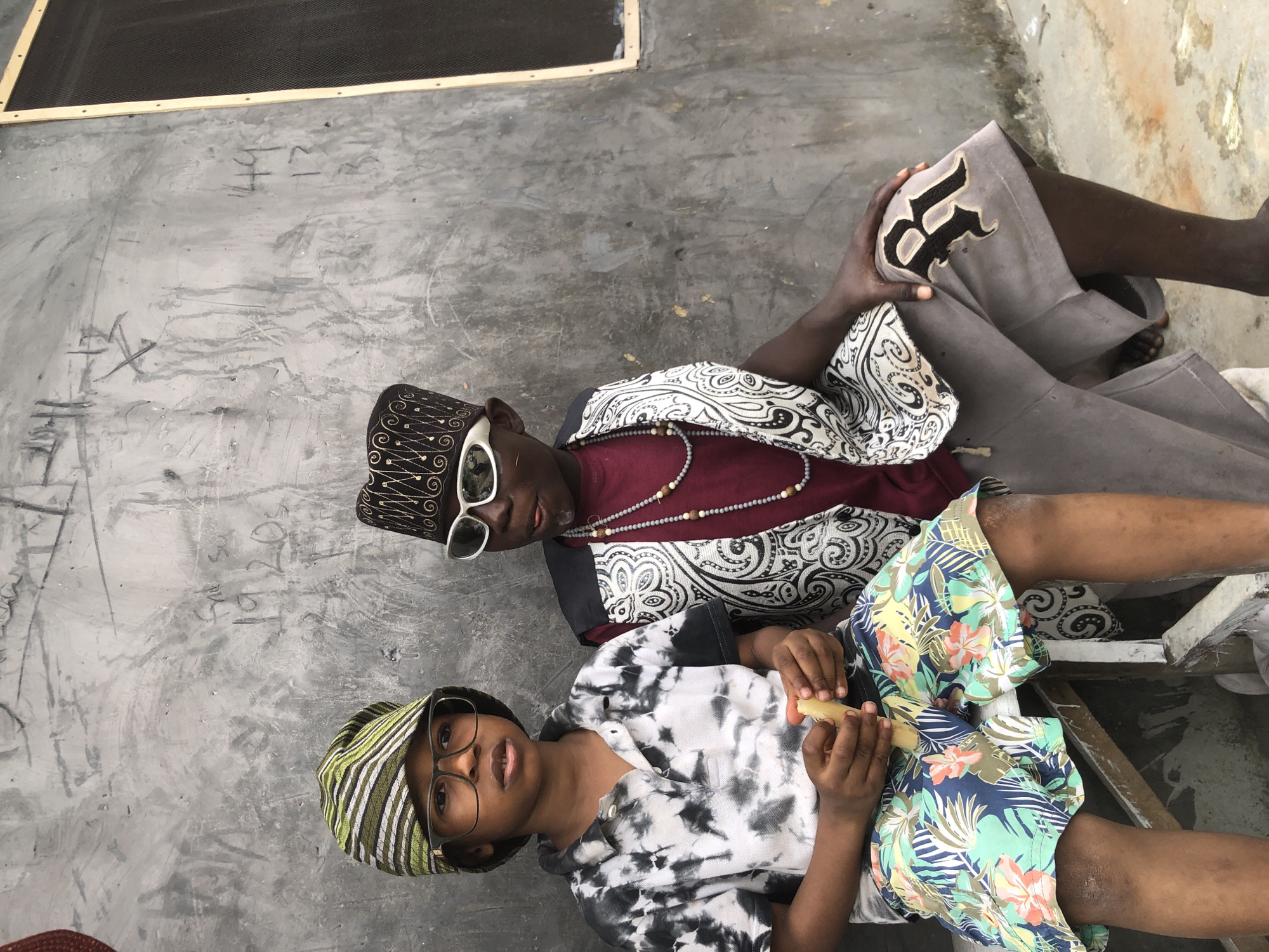 Two young boys are seated on a wooden bench against a rough, concrete wall in Ado, Nigeria. The boy on the left wears a tie-dye polo shirt and floral shorts, a green and yellow striped cap, and glasses. He holds a piece of food in his hands. The boy on the right wears a maroon shirt, a patterned vest, a black and gold embroidered cap, and white sunglasses. He has beads around his neck and is wearing grey shorts with an embroidered "R" on the left leg. The wall behind them is covered in graffiti and markings, including numbers that appear to read "1920s" and "144" and "13". A screened window is visible on the far right side of the wall. The ground is rough concrete.
