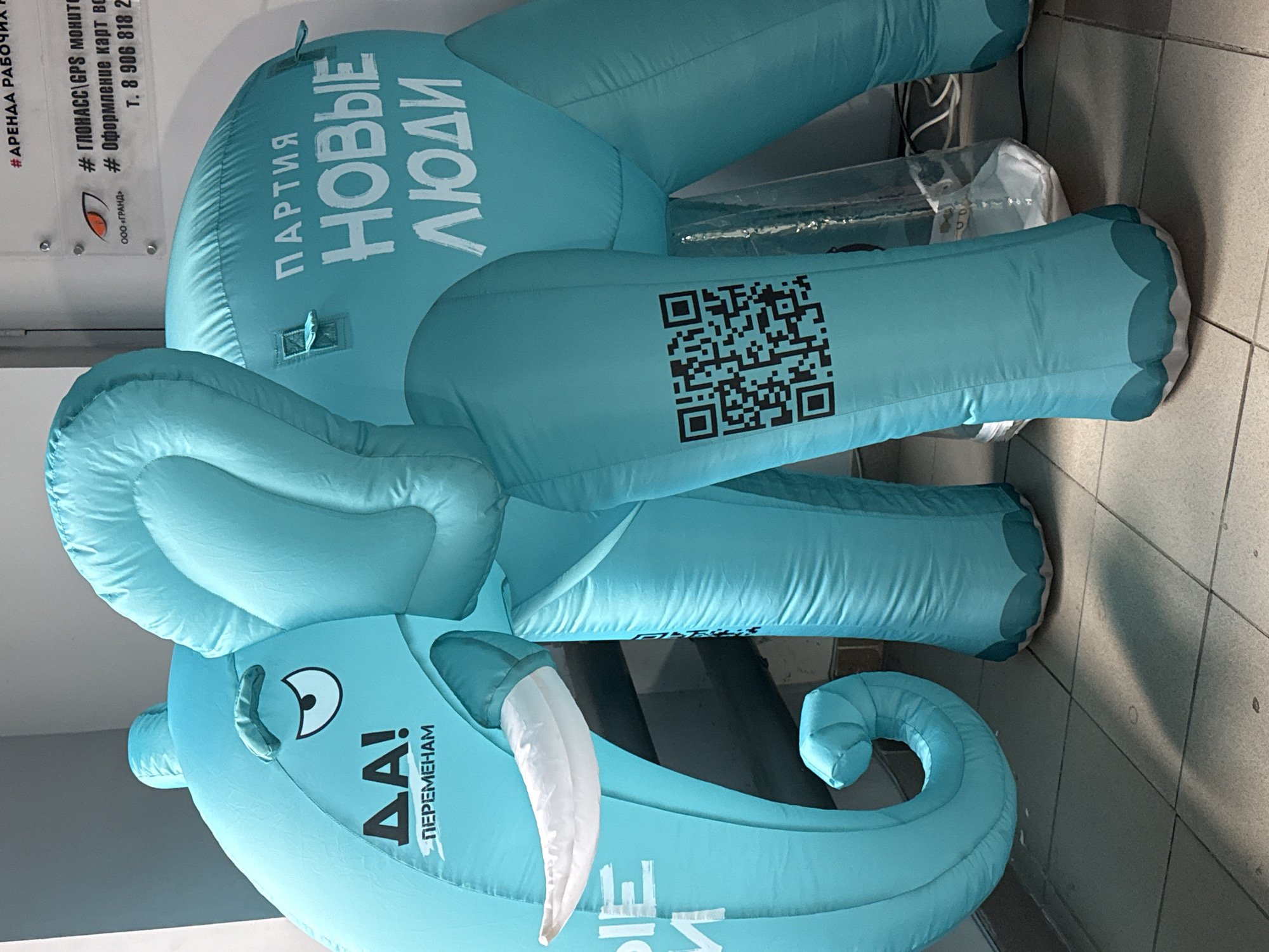 An indoor shot captures a large, light blue inflatable elephant serving as a promotional display in what appears to be a commercial or public setting in Votkinsk, Russia. The elephant, depicted with a friendly cartoon eye, white tusks, and a curled trunk, bears political slogans and a QR code. On its side, the text "ПАРТИЯ НОВЫЕ ЛЮДИ" (Party New People) is prominently displayed, identifying it as a symbol for the Russian political party. On its head, the phrase "ΔA! ПЕРЕМЕНАМ" (YES! FOR CHANGES) conveys an affirmative message. A black QR code is visible on one of its legs, likely leading to more information about the party. The elephant stands on a grey tiled floor, adjacent to a white wall adorned with commercial signs. These signs, partially visible, advertise services such as "#АРЕНДА РАБОЧИХ" (RENT WORKERS), "# ГЛОНАСС/GPS МОНИТОРИНГ" (GLONASS/GPS MONITORING), and "Оформление карт водителя" (Driver card processing) from "ООО «ГРАНД»" (LLC "GRAND"), along with a partial phone number. The scene suggests a static, attention-grabbing display intended for public engagement, possibly within a retail space, exhibition hall, or a public office.