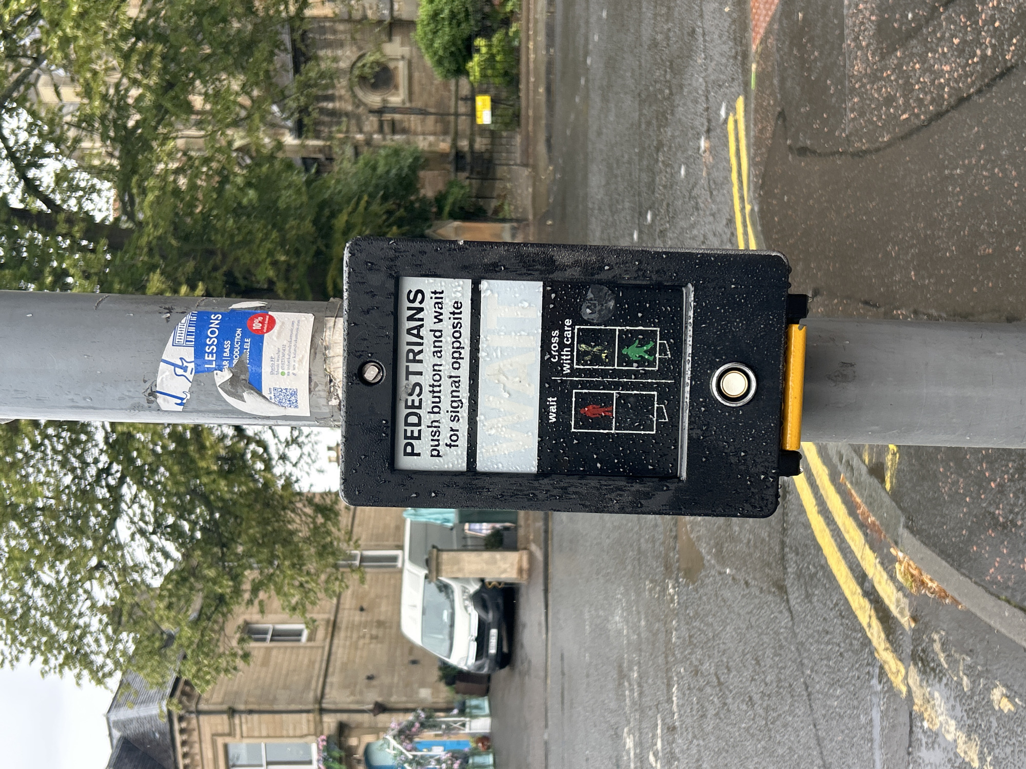 An eye-level medium shot captures a pedestrian crossing signal box on a wet, overcast day in the City of Edinburgh, United Kingdom. The black signal box, speckled with raindrops, is mounted on a grey metal pole. The box displays instructions for pedestrians: "PEDESTRIANS push button and wait for signal opposite". Below this, two panels indicate "wait" with a red standing figure and "cross with care" with a green walking figure. A circular white button is located at the bottom of the box, above a bright yellow trim. The background features a wet road with double yellow lines, a stone building with a grey tiled roof, trees with lush green foliage, and a white van parked on the street. The overall impression is of a typical urban scene, made more somber by the damp weather.