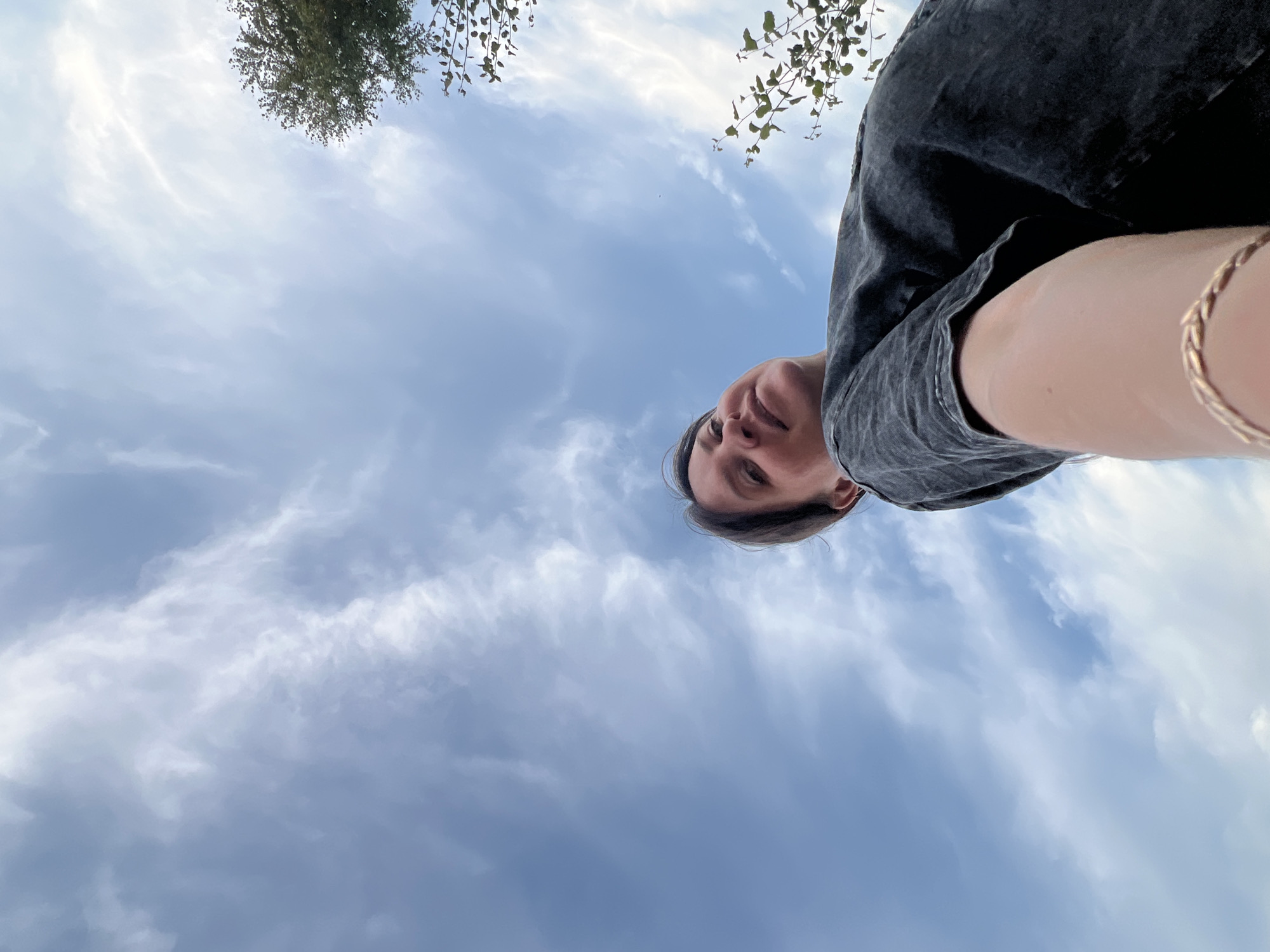 The media file is a low-angle, upside-down selfie taken outdoors. The subject is a person whose head and upper torso are visible, looking up towards the camera. They are wearing a dark gray t-shirt. A thin, gold-colored bracelet is visible on their left wrist.
The background is predominantly a blue sky with scattered white clouds, suggesting it is daytime. There are also glimpses of green foliage from trees in the upper left and upper right corners of the image. The perspective suggests the photo was taken on a clear day, possibly with some wispy clouds. There is no visible text in the image. The overall mood appears casual and relaxed.