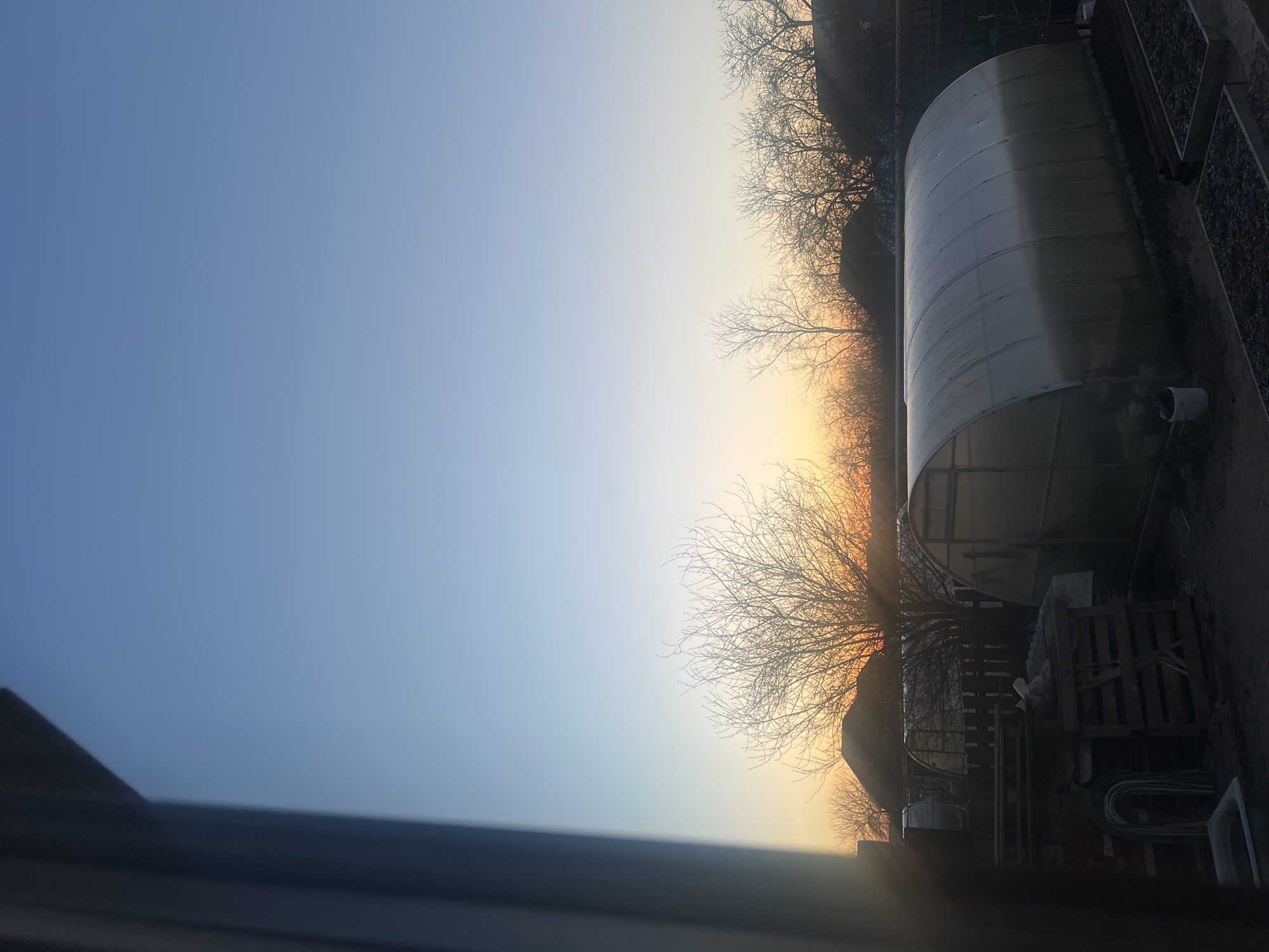 The image shows an outdoor scene at sunrise or sunset. The sky transitions from a blue hue to an orange glow near the horizon. Bare trees are silhouetted against the sky. A large, cylindrical structure is present, potentially a greenhouse. Various objects such as pallets, stacked materials, hoses, and a white sink are visible. There is no verifiable information indicating that this scene is located in Votkinsk, Russia. No people are present in the image.