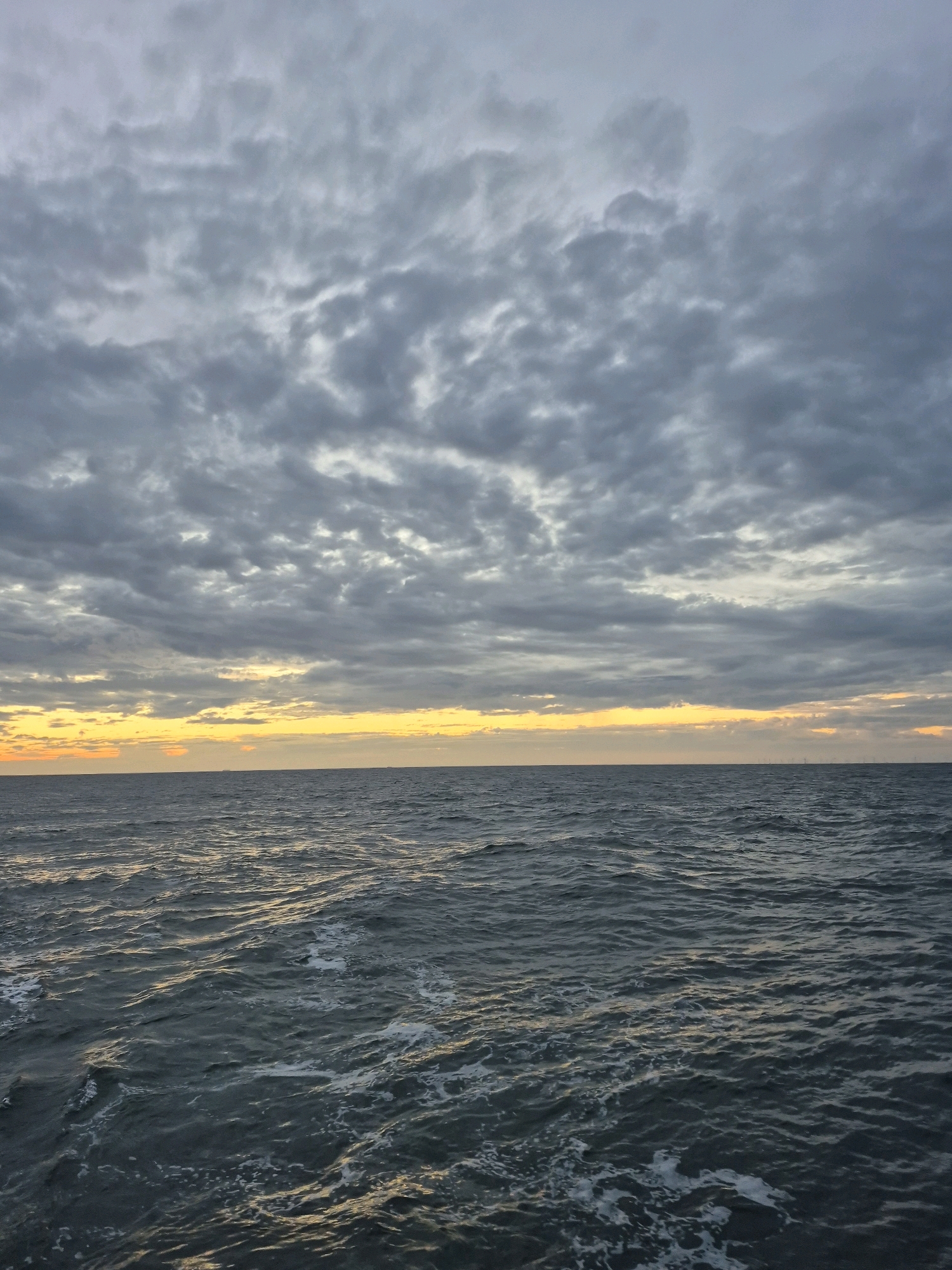 The image depicts a body of water under a cloudy sky, likely the sea given the horizon. The water is a dark, choppy gray-blue with white foam visible on the surface, suggesting movement. The sky is filled with dense, overcast clouds in varying shades of gray, with a band of lighter, yellowish-orange light peeking through near the horizon, indicating either sunrise or sunset. The horizon is a sharp line separating the sea from the sky, with faint, distant shapes that could be ships or structures barely visible. There are no people or animals in the frame. The scene is captured from a low vantage point, looking out over the water. The location is identified as Unknown, Germany.