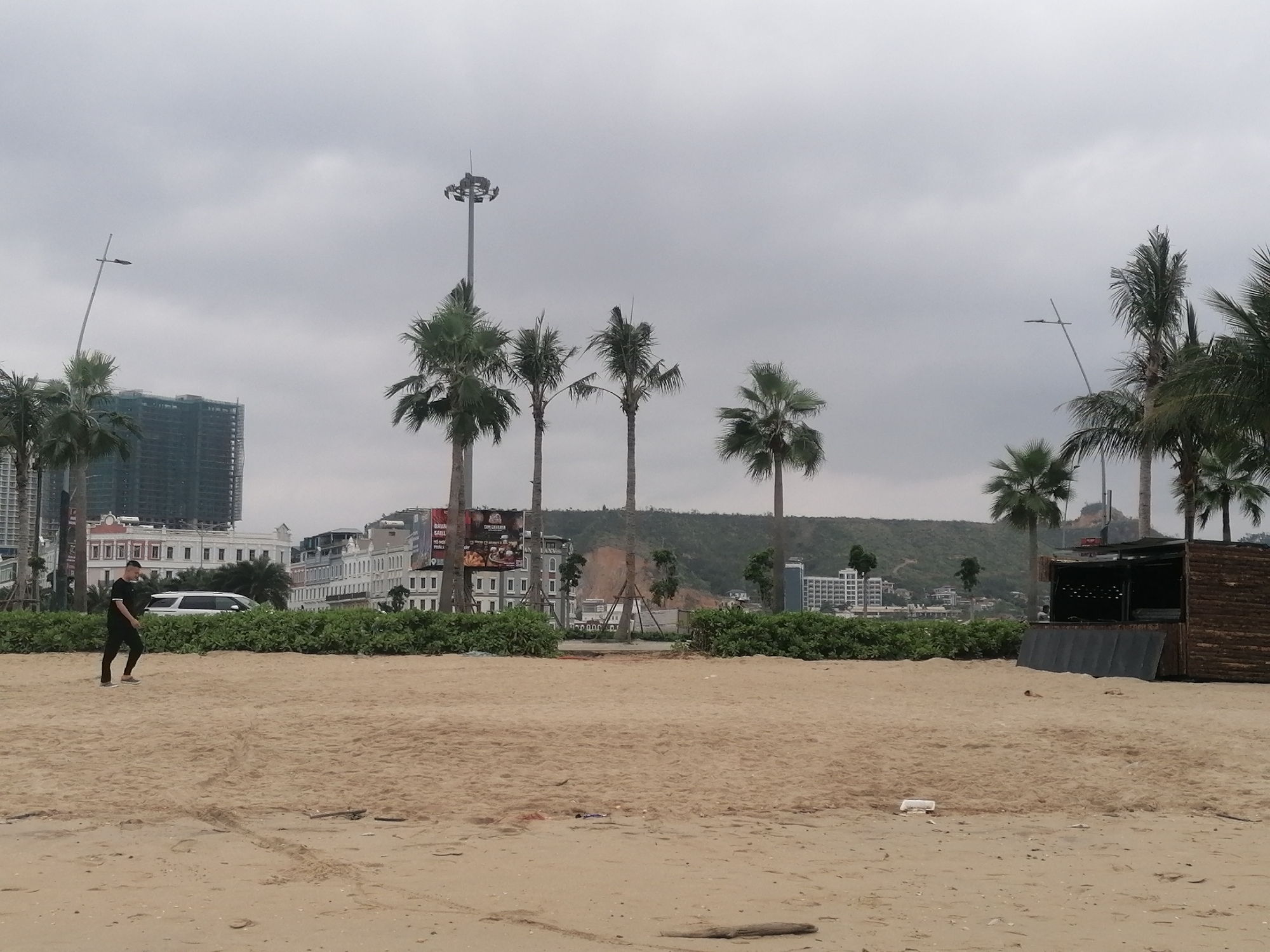 The image presents a wide view of a beach scene in an urban coastal environment under an overcast sky. In the foreground, expansive light-brown sand stretches across the frame, showing faint tracks and minimal debris. A man, wearing a black t-shirt, dark pants, and light-colored shoes, walks from left to right across the sandy area, looking downwards or straight ahead.
The mid-ground features a row of vibrant green bushes separating the sandy beach from the built environment. Several tall palm trees are scattered throughout, their fronds reaching towards the cloudy sky. A prominent, very tall metal light mast with multiple floodlights stands near the center. To the right, a rustic wooden kiosk or bar structure, appearing closed, sits on the sand, with bottles visible on interior shelves.
Beyond the trees and bushes, a mix of buildings forms the background. On the left, modern high-rise structures, one partially obscured by scaffolding, are visible. In the center, a series of classical-style white buildings with red roofs line a street, and a white SUV is parked nearby. A large billboard above these buildings displays partially legible text, including "BANH MI SAUL" and "COFFEE." Further back, green hills rise, with sections of exposed reddish-brown earth suggesting recent land alteration.
The weather is overcast with a uniformly gray, cloudy sky, indicating a diffused light and possibly a mild, humid atmosphere during the daytime. The blend of sandy beach, palm trees, urban architecture, and natural hills provides strong location cues consistent with a developed coastal area. No explicit emotions are conveyed, and the scene appears calm.