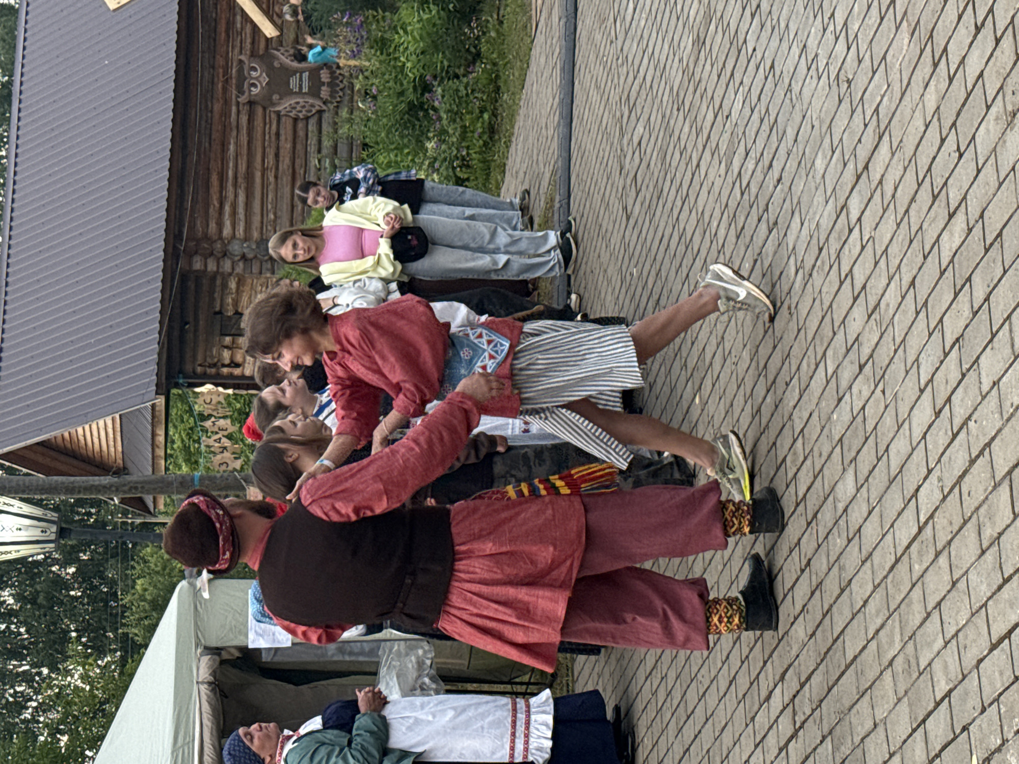 The image shows a group of people outdoors, possibly at a festival or gathering, in Старые Быги, Russia. The setting includes a rustic wooden building with a metal roof on the left and a paved walkway made of grey bricks. Lush green vegetation with purple flowers is visible in the background.
Several individuals are featured, dressed in a mix of casual and traditional attire. One woman in the foreground, wearing a red top and a dark vest over a patterned skirt and maroon pants, appears to be dancing or performing. She has a patterned headband and a brown hat. Next to her, another person in a red top and striped skirt is also engaged in movement. Further back, a group of women are standing, dressed in modern clothing, including a woman in a yellow cardigan over a pink top and light blue jeans. A man in a blue plaid shirt is also visible.
The scene suggests an outdoor event taking place during the day, likely with pleasant weather, given the presence of many people outdoors. The attire of some individuals, particularly the woman in the red and maroon outfit, hints at a cultural or folk performance. The overall mood appears lively and engaged. No visible text is clearly legible in the image.