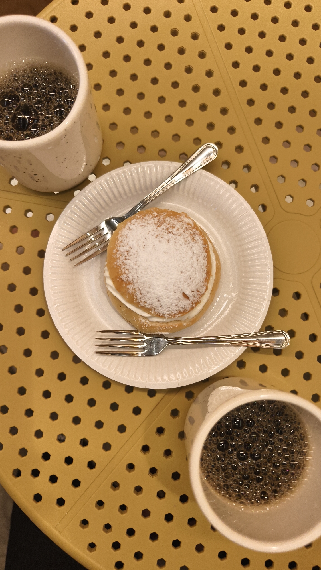 A high-angle shot depicts a snack and beverage setting on a yellow, perforated table. Two white mugs filled with dark liquid, possibly coffee, are positioned on the table's periphery. Centrally located is a white, ridged paper plate holding a round pastry topped with powdered sugar and filled with a creamy substance. Two silver forks are placed on the plate, one above and one below the pastry. The location is possibly in the city of Tyumen, Russia. No people or actions are visible in the image.