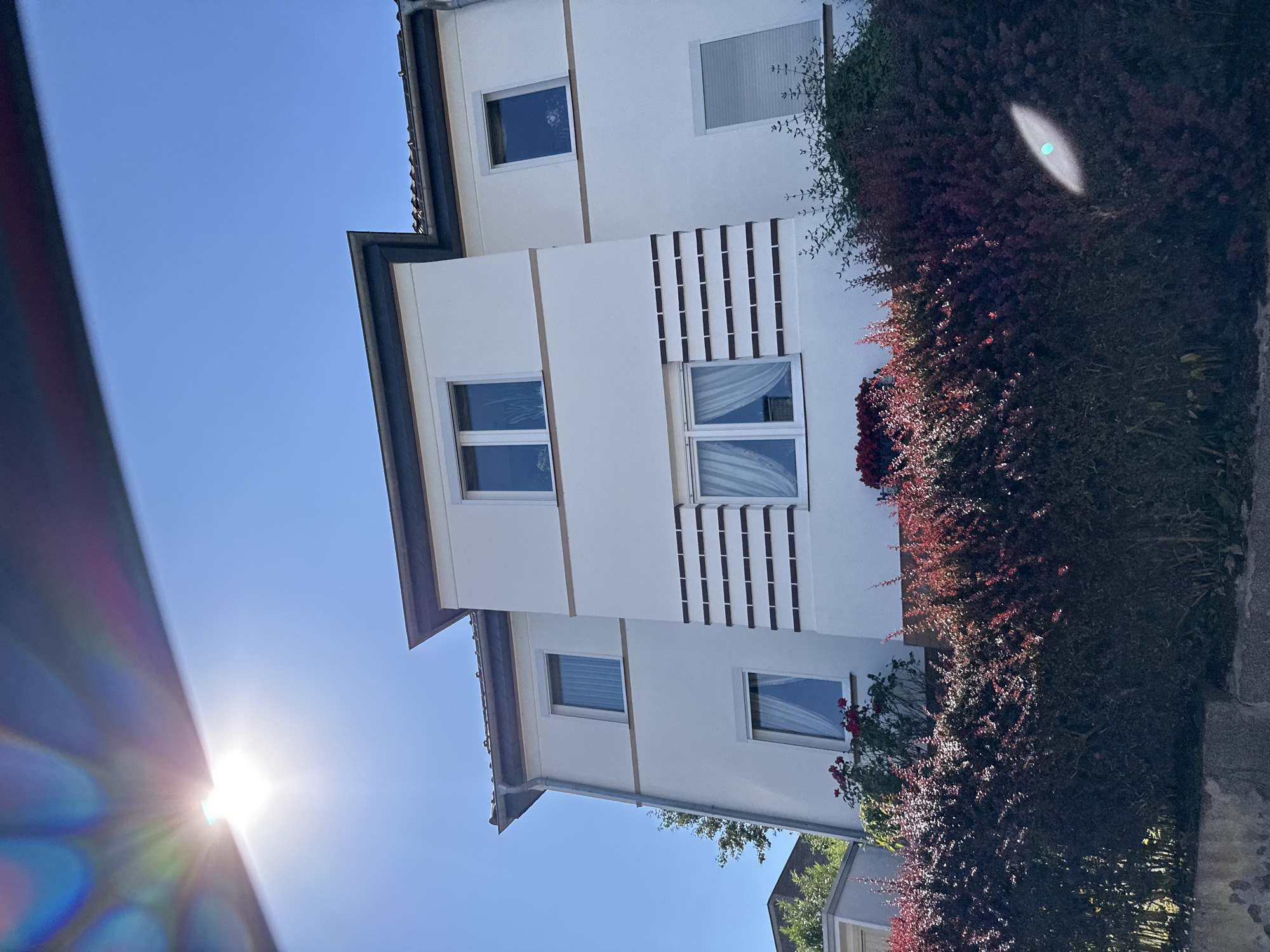 The image shows a modern white apartment building with a dark-rimmed roof, set against a clear blue sky. The building features several windows, some with white curtains. A distinctive decorative element of horizontal dark brown rectangles is present on the facade. In the foreground, a dense, dark red-leaved bush lines the front of the property. A bright sun glare is visible in the upper left corner of the image, and a smaller lens flare appears in the lower right. The architecture suggests a residential area in Bad Sachsa, Germany.
