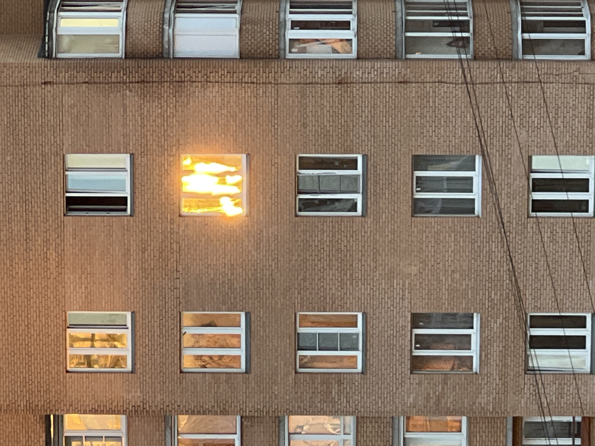 The image shows a brick apartment building in Sinyavino, Russia, at dusk. Several windows are visible, some with reflections of the setting sun. One window on the third floor is illuminated by a bright orange and yellow light, suggesting the presence of fire. The other windows are dark or show faint interior lighting. Electrical wires are visible across the facade of the building.