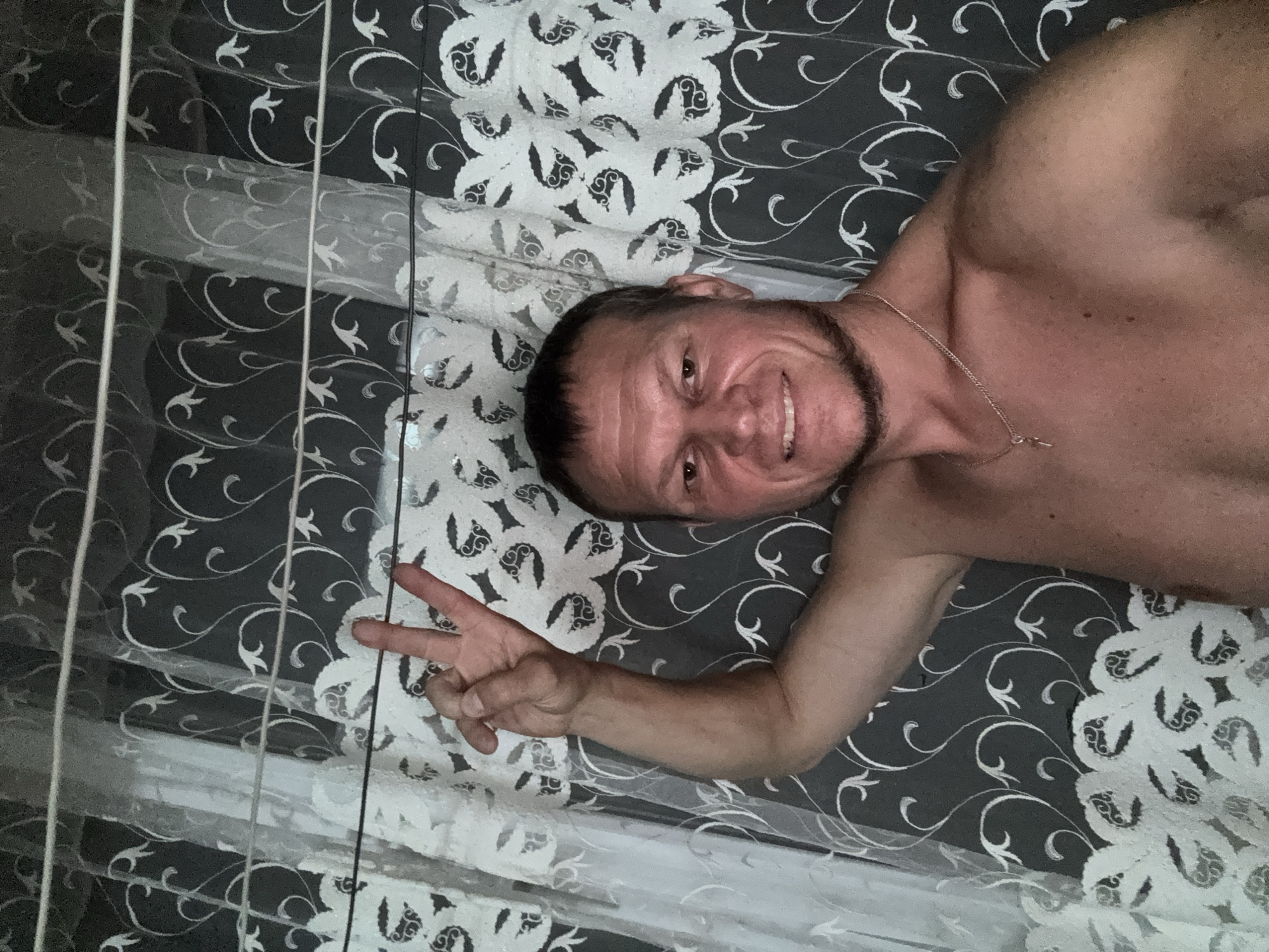 The image shows a shirtless man with short brown hair and a goatee, smiling and making a peace sign with his right hand. He is wearing a silver chain with a cross pendant. The man is standing in front of a dark curtain with a white floral pattern, and a lighter sheer curtain is visible to his left. The background suggests an indoor setting, possibly a bedroom or living area. There are clotheslines or wires running horizontally across the top of the image. The man's skin appears flushed, suggesting warm lighting or an elevated body temperature. The overall impression is a casual selfie taken indoors.