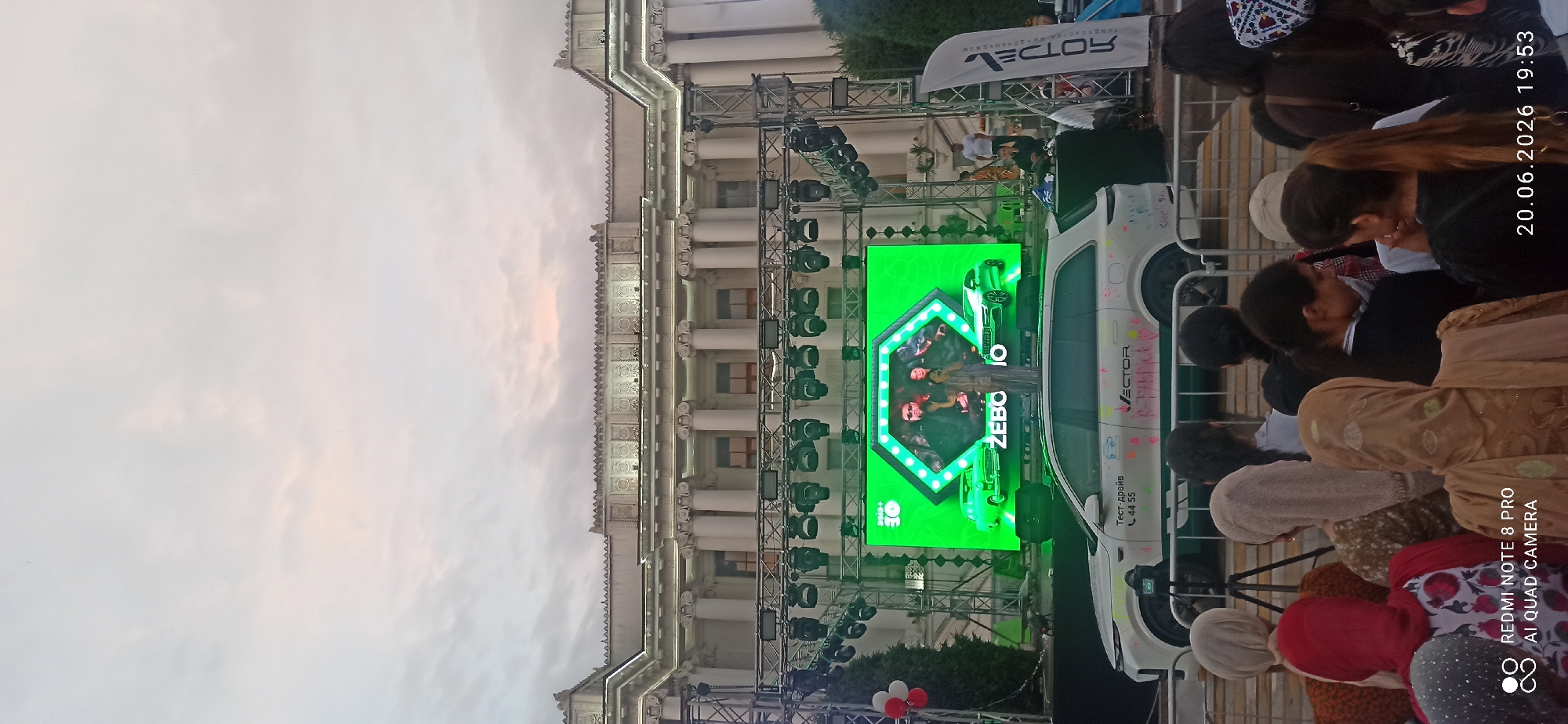 In an outdoor urban setting, a stage is set up in front of a grand building with classical architecture. A large LED screen displays an image of two individuals and two cars. A white SUV is parked in front of the stage, bearing markings and text including "Тест-драйв" and "VECTOR". A crowd of people, many wearing headscarves, are gathered in front of a barrier, observing the event. The sky is overcast with some soft pink and orange hues suggesting either dawn or dusk. A camera on a tripod is positioned in front of the SUV. A tall, vertical banner with text is visible on the right side of the frame. The overall scene suggests an outdoor event or promotion.