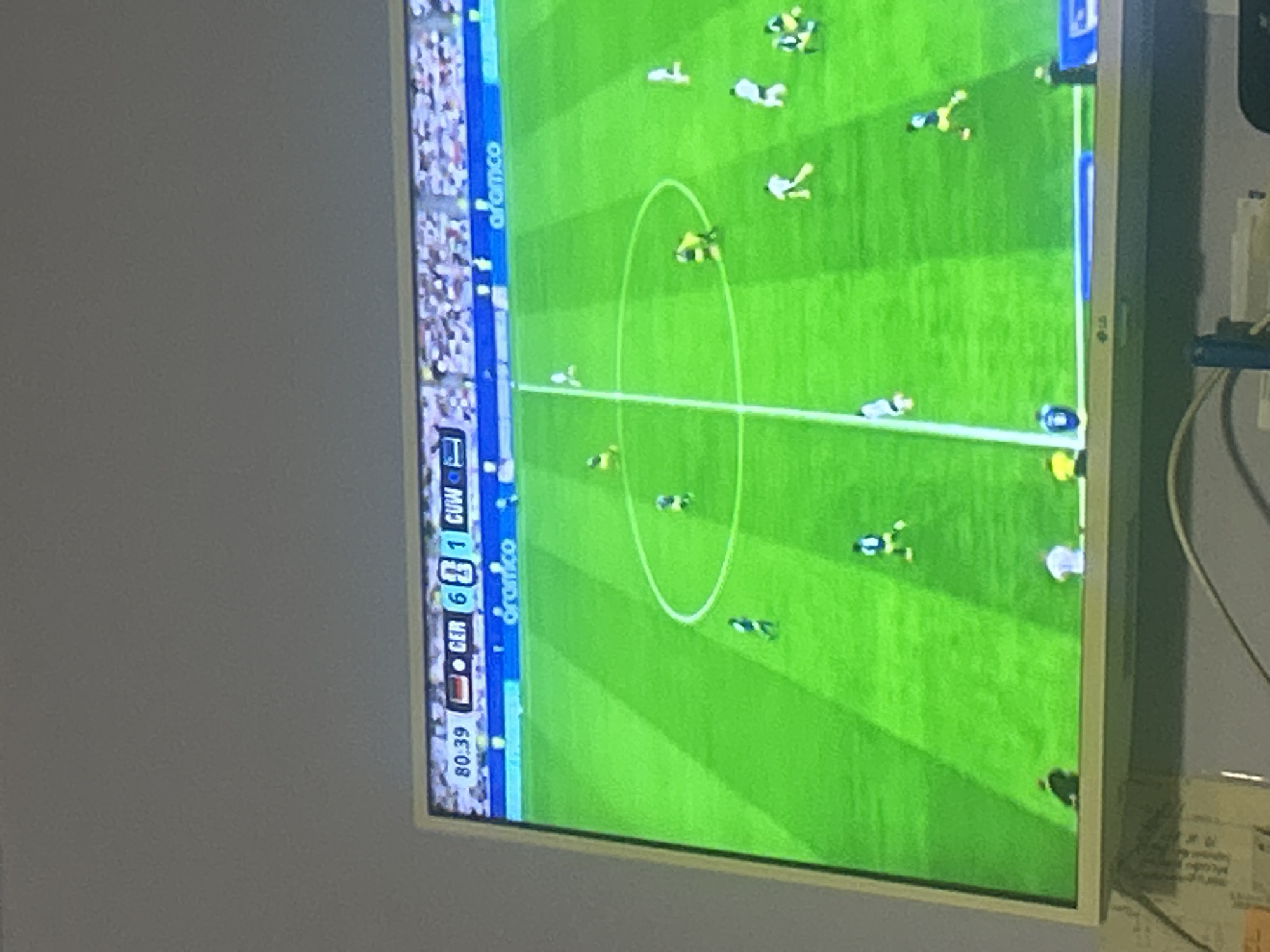 The image shows a television screen displaying a soccer match. The score is Germany 6, Kuwait 1, with the game at 80:39 minutes. The field is green with white lines. Several players in yellow and white uniforms are visible on the field. The word "aramco" is displayed on a banner in the background. The bottom of the image shows the lower part of the television frame and some objects below it, including cables and a wall. The location is not specified, but it appears to be an indoor setting where the television is placed.