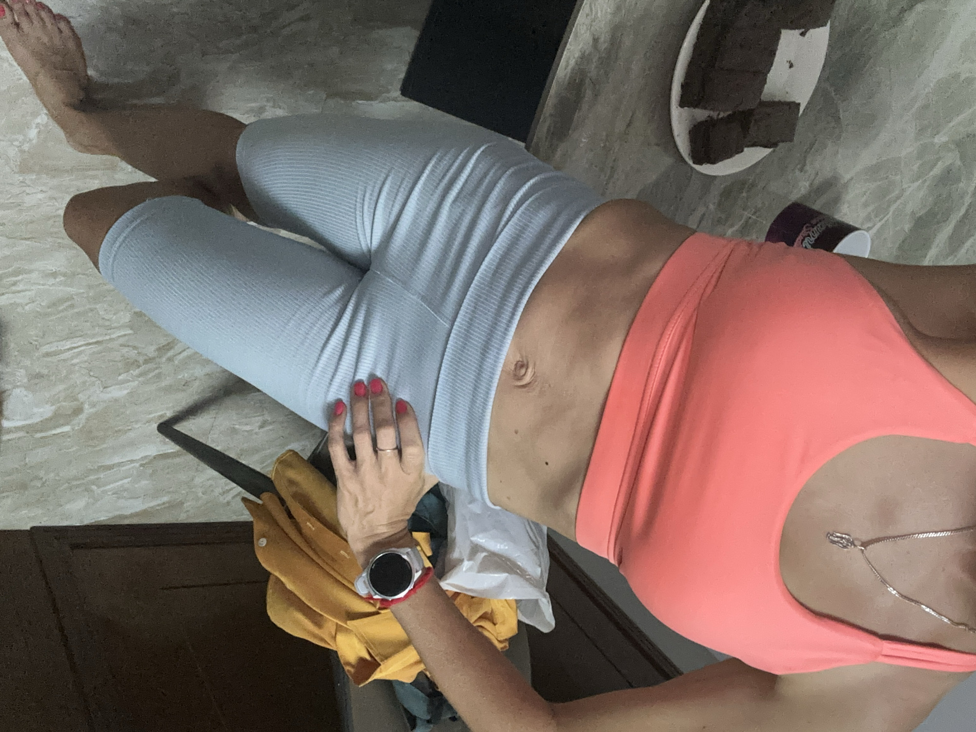 A woman lies on a marbled floor, wearing light blue ribbed leggings and a coral-colored sports bra. Her right foot is visible at the top left corner of the frame. A silver necklace hangs around her neck. Her left hand, with red nail polish, rests on the leggings, wearing a wedding ring on her ring finger and a smartwatch on her wrist. A yellow shirt is draped to the lower left of her hand. In the upper right corner, there's a plate of dark-colored square food items, a dark cup, and a dark rectangular object. The location is within a building or apartment in Bac Nha Trang Ward, Vietnam.