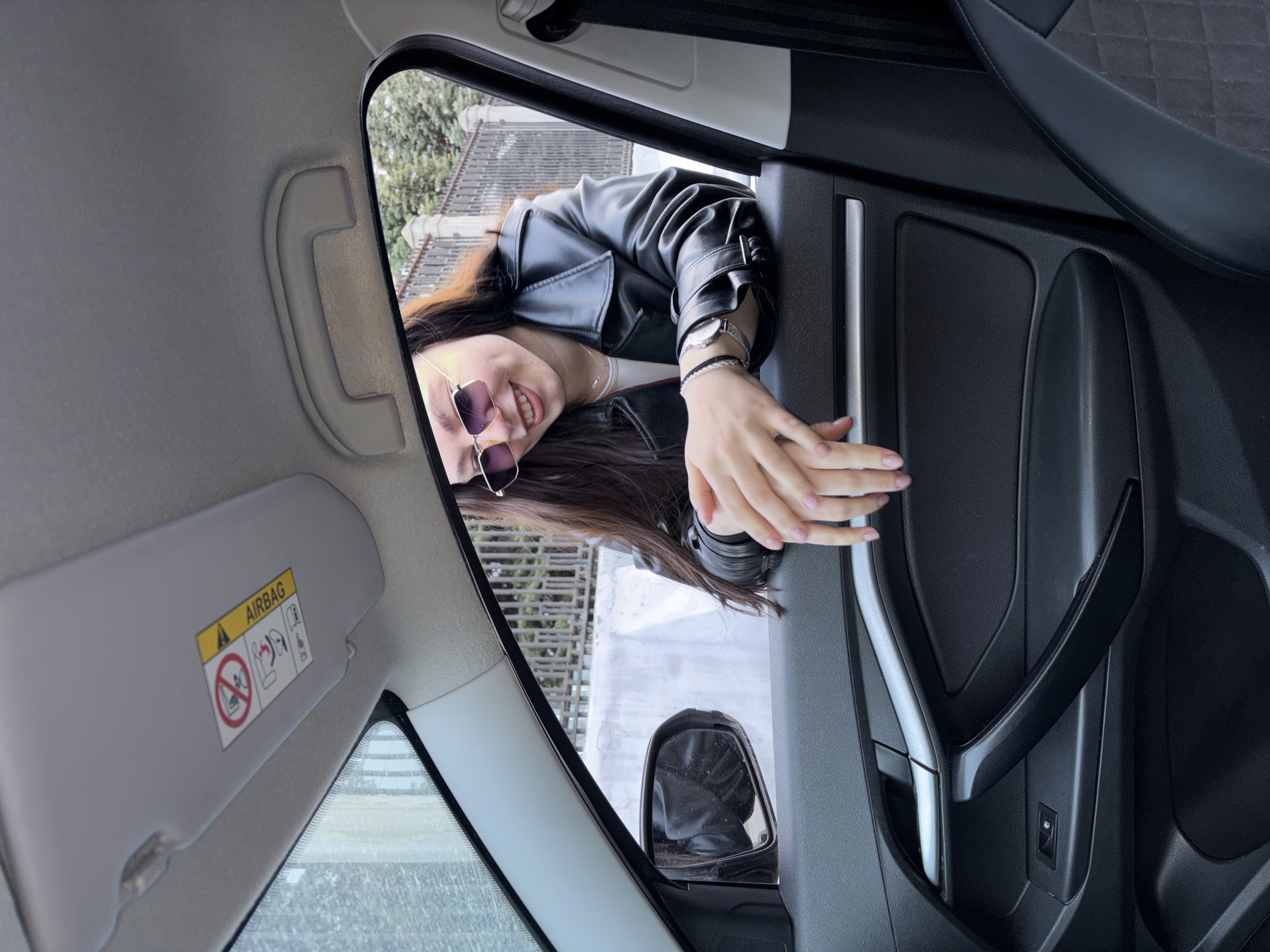 A young woman is photographed from within a car, appearing to lean into the open window. She wears sunglasses and a black leather jacket and is smiling slightly. Her wrists are adorned with a bracelet and a watch. The interior of the car is visible, including the door panel, a sun visor labeled "AIRBAG", and a grab handle. The rearview mirror is also visible through the open window. The background visible through the window appears to be an urban setting with a wall or fence. There is no verifiable data regarding the location as the city of Perm, Russia.