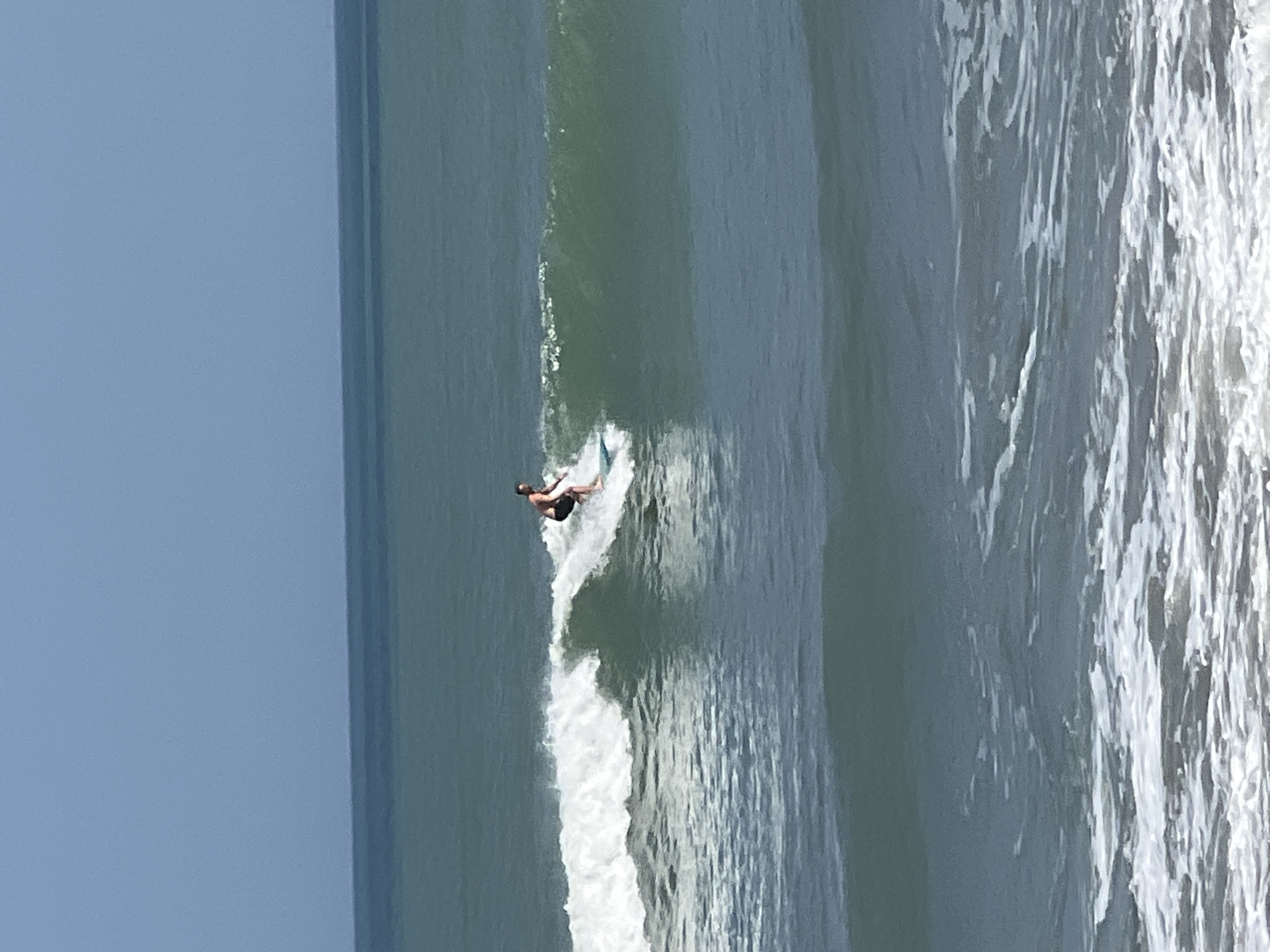 A lone surfer rides a breaking wave off the coast of Kuta, Indonesia. The surfer, a man with a dark complexion and wearing black shorts, is positioned on a light-colored surfboard. He is mid-action, leaning slightly to his right, as he glides down the face of the wave. The wave itself is a cresting, whitewash of foam that is breaking to the left of the surfer. The ocean water is a deep teal color, with lighter blues and greens visible in the distance, suggesting a clear day. The sky above is a pale, hazy blue. The scene is captured from a medium distance, showing the surfer and wave clearly against the backdrop of the ocean and sky.