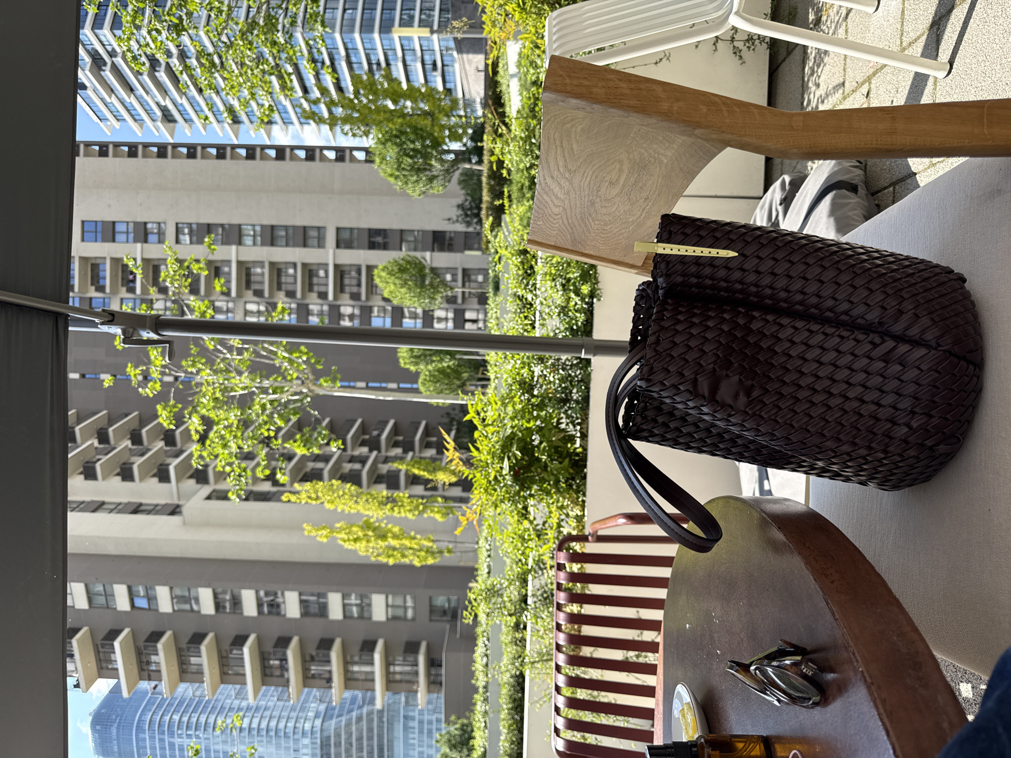 A view from an outdoor cafe or patio, with modern buildings of Belgrade, Serbia in the background. The foreground features a table and chair, with a brown woven leather handbag on the chair. There are some personal items on the table, including what appears to be a pair of glasses and a small bottle. The environment consists of green trees, plants, and urban infrastructure.