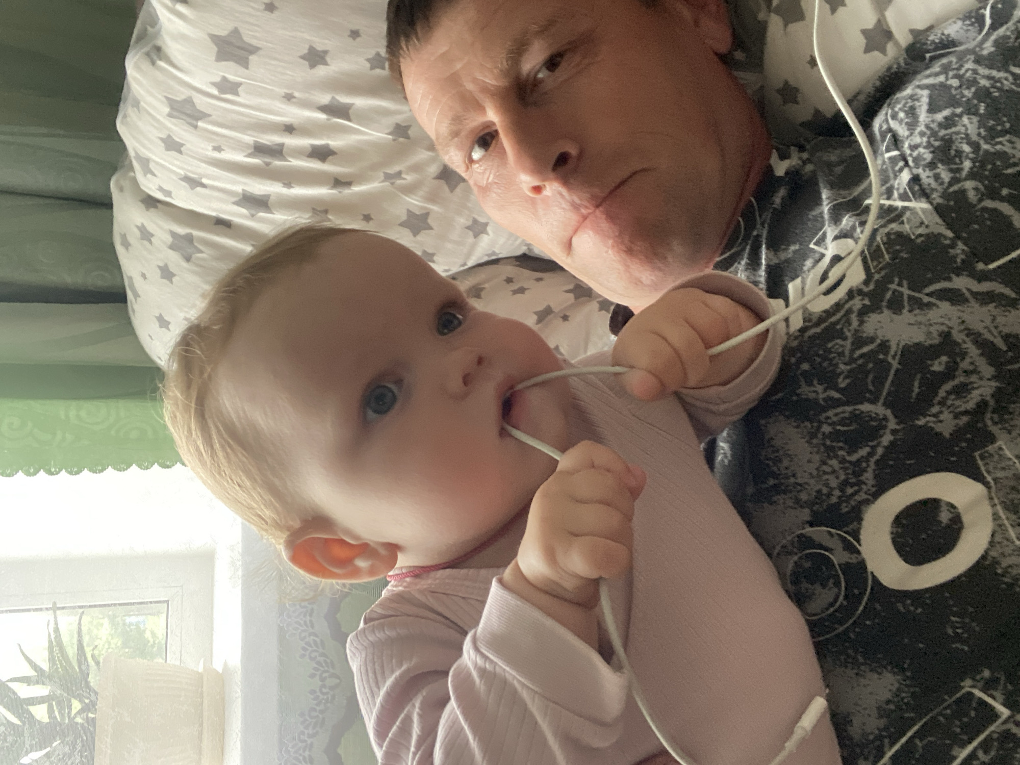 The image captures an overhead view of an adult male and a baby lying down indoors, likely in a bedroom or living area. The adult, positioned on the right, has short dark hair and looks directly towards the camera with a neutral to slightly serious expression. He is wearing a dark-colored long-sleeved shirt with abstract white or light grey patterns, including a distinct white circular shape resembling the letter "O" on the chest area. He is resting his head on a light-colored pillow adorned with grey star patterns.
To the left and slightly below the adult, a baby or toddler is visible, lying on their back with their head turned towards the left. The baby has light blonde hair, blue eyes, and is wearing a light pink long-sleeved top. They are actively engaged with a white earbud wire, holding it with both hands and seemingly putting part of it into their mouth.
The setting appears well-lit by natural light streaming in from a window visible in the background to the bottom left. A green scalloped curtain hangs above the window, through which green foliage can be seen, suggesting an outdoor view. A white, possibly decorative, object stands near the window. The overall scene suggests a relaxed daytime moment of interaction, possibly in a home in Фоки, Russia.
