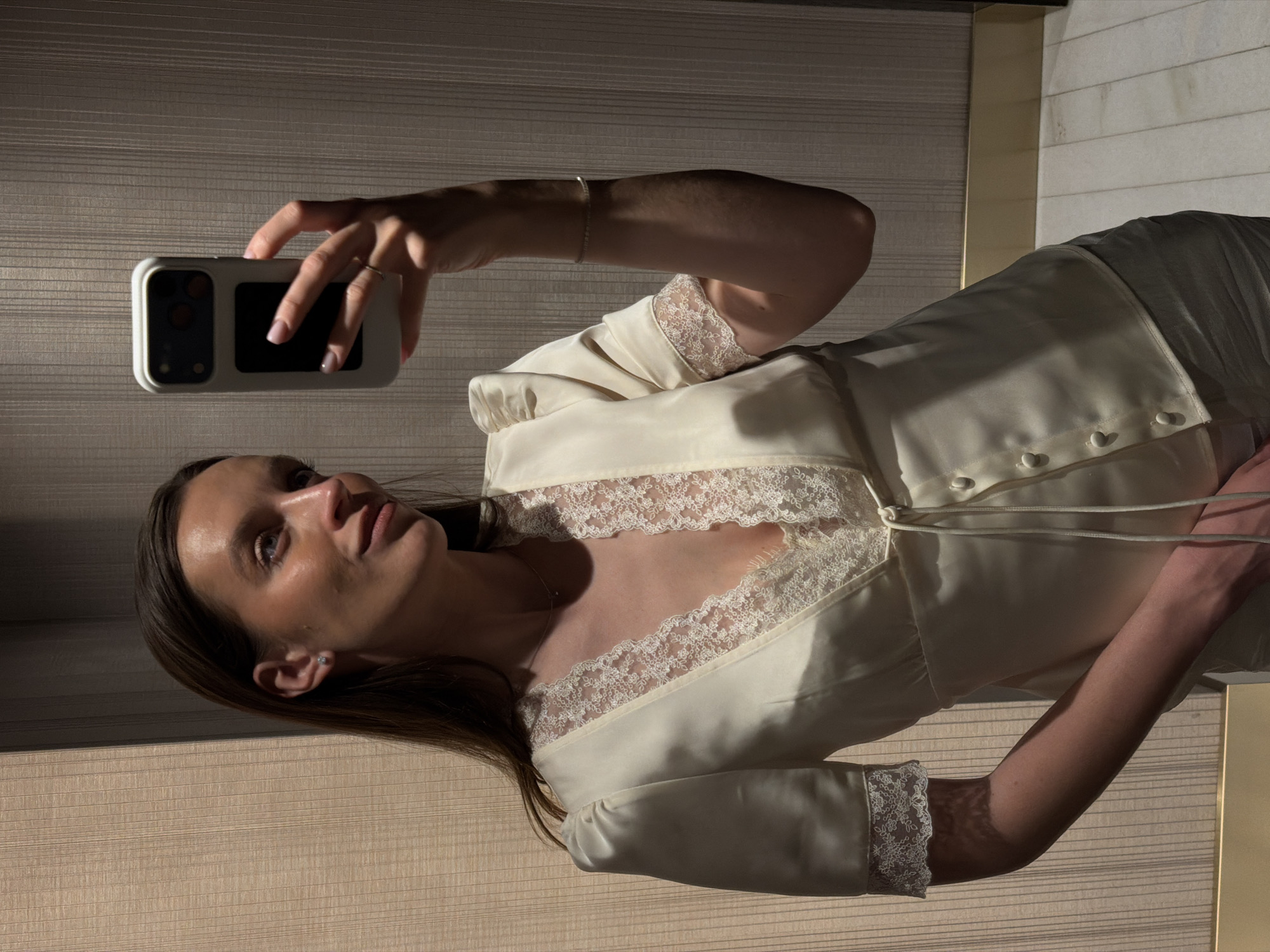 A woman is taking a selfie in a location that is difficult to pinpoint to Budva, Montenegro, from the image alone. She is holding a cell phone up with her left hand. She has light brown hair and is wearing a light-colored top with lace detailing at the neckline and sleeves. Her skin appears fair, and she has a stud earring in her left ear. The background includes a textured wall that is horizontally oriented and two columns.