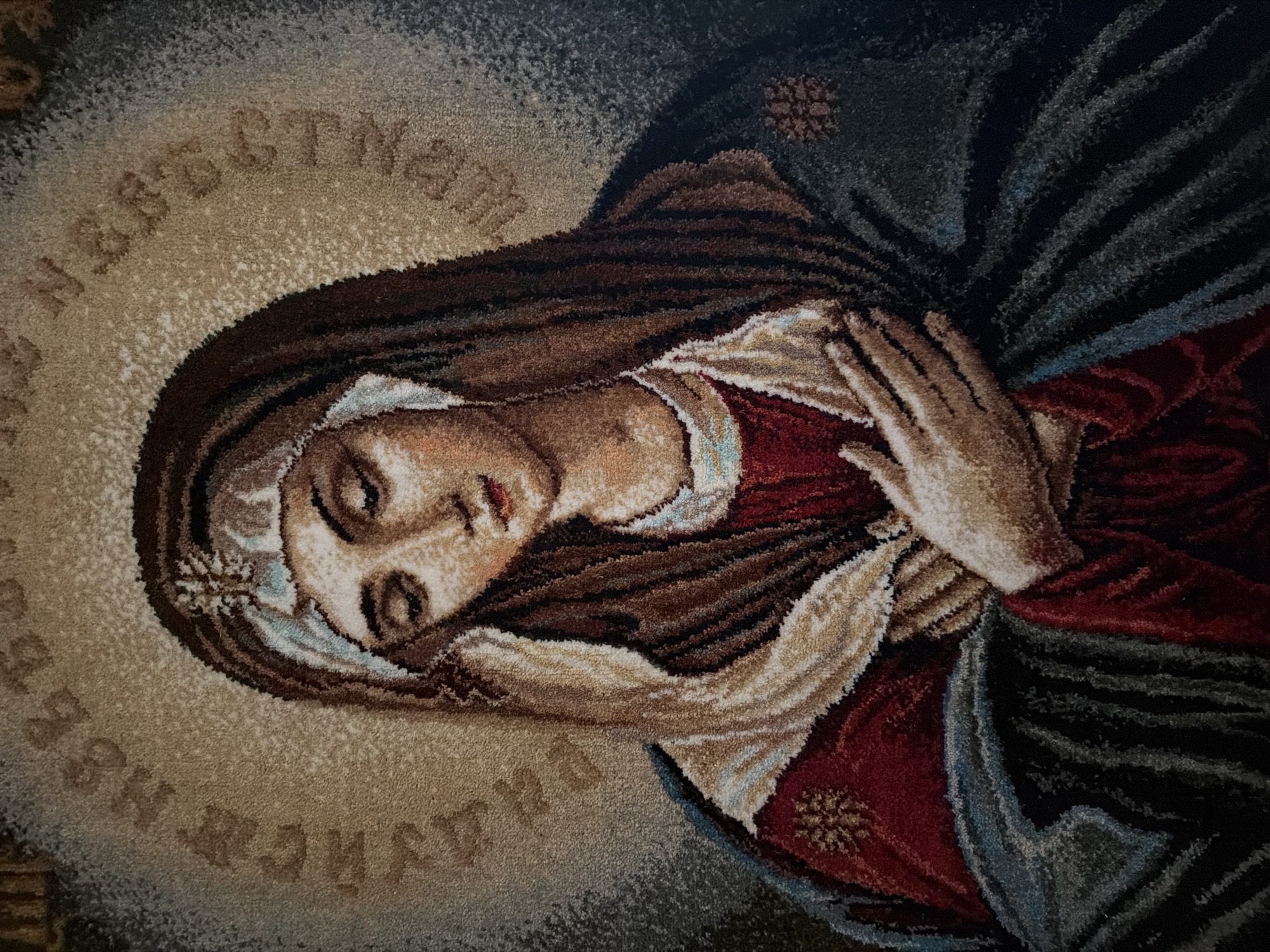 The image depicts a rug or tapestry featuring a portrait of the Virgin Mary. Her face is pale, with closed eyes and a serene expression. She is depicted wearing a dark brown head covering with a lighter-colored trim and a small, flower-like decoration. She is clad in a red garment, draped with a beige cloth and a dark blue cloak. Her hands are clasped together in prayer. The portrait is surrounded by a circular halo of lighter shades, containing text in a Slavic language. The background is a mix of dark and light shades, suggesting a neutral setting. There is no information to suggest this is in the city of Levkovo, Russia.