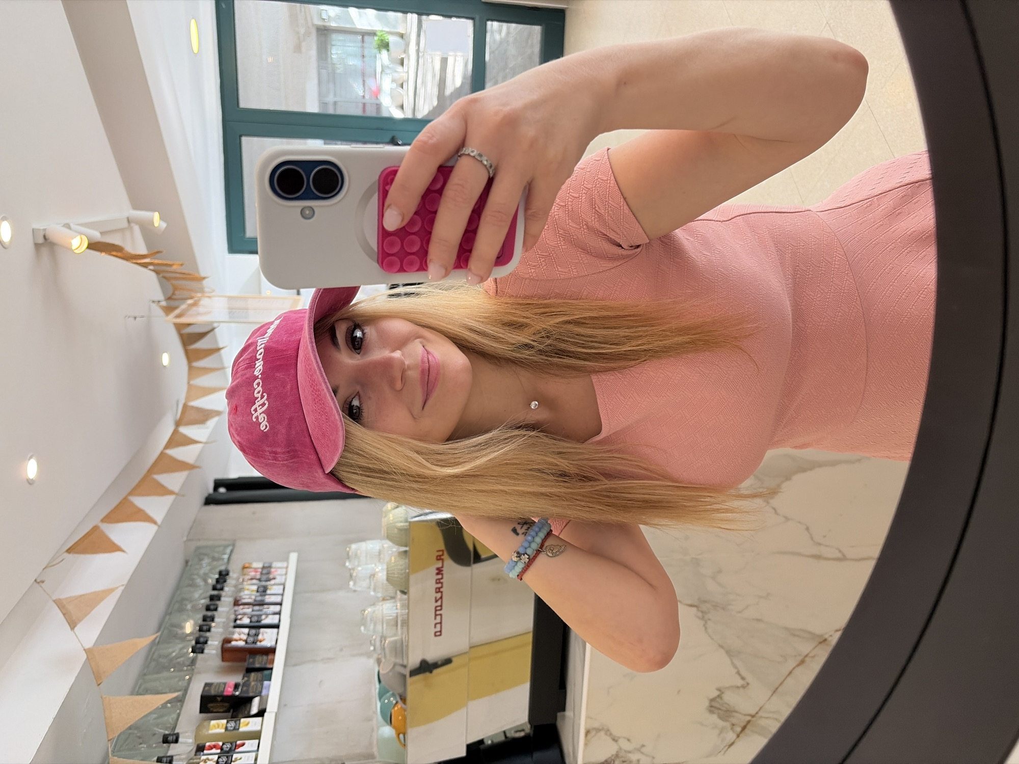 A young woman with long blonde hair is taking a selfie in a mirror. She is wearing a pink cap and a pink dress. She is holding a smartphone with a white case and pink pop sockets. She also wears a ring and bracelet with charms. The background shows a café or shop interior with a display of products on shelves and a coffee machine. Pennant banners are visible along the ceiling, suggesting a festive or decorative environment. The location seems to be in a well-lit indoor space. A window or door frame is visible in the background, possibly suggesting a location in the city of Budva, Montenegro based on prior information.